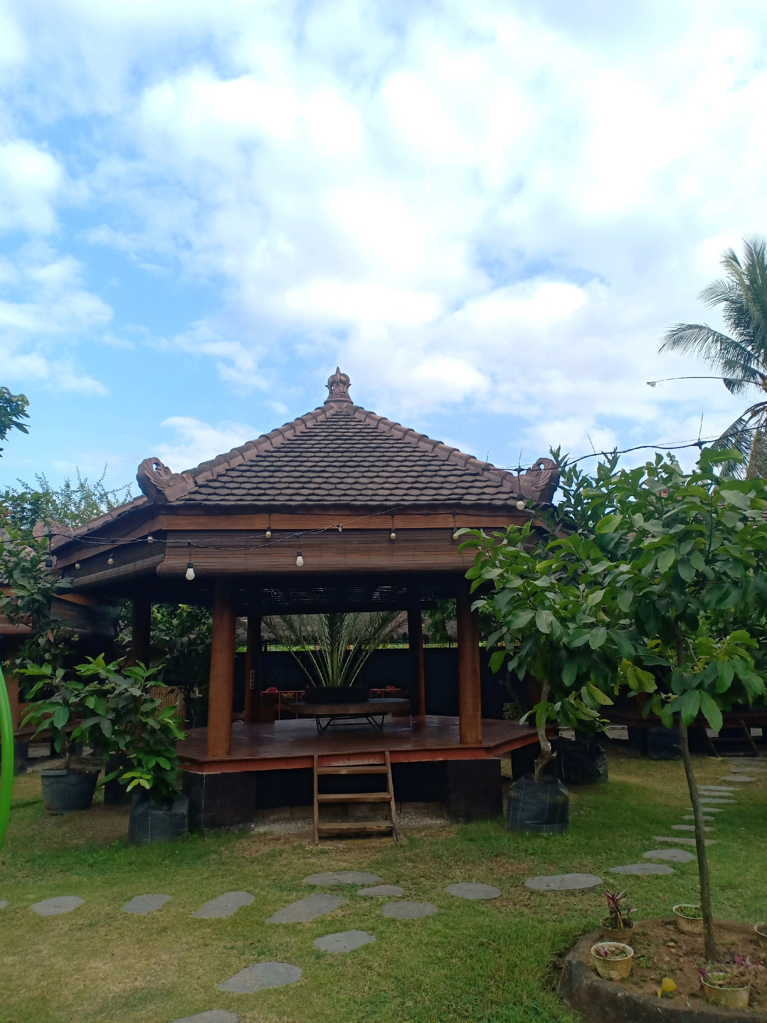 The image depicts a traditional Balinese-style gazebo set within a garden. The gazebo features a tiered, tiled roof with ornate decorative elements on the eaves. Wooden pillars support the roof, and the structure is elevated on a wooden platform. Inside the gazebo, a wooden table and benches are visible. The surrounding environment is lush and green, with a well-maintained lawn and various trees and potted plants. Stone stepping stones are laid out on the grass, leading towards the gazebo. In the background, a palm tree and a bright blue sky with scattered white clouds suggest a tropical or subtropical climate. The scene is tranquil and inviting, suggesting a peaceful outdoor seating area, possibly in a resort or restaurant. This location is identified as being in the city of Langkap, Indonesia. There are no people or animals present in the image.