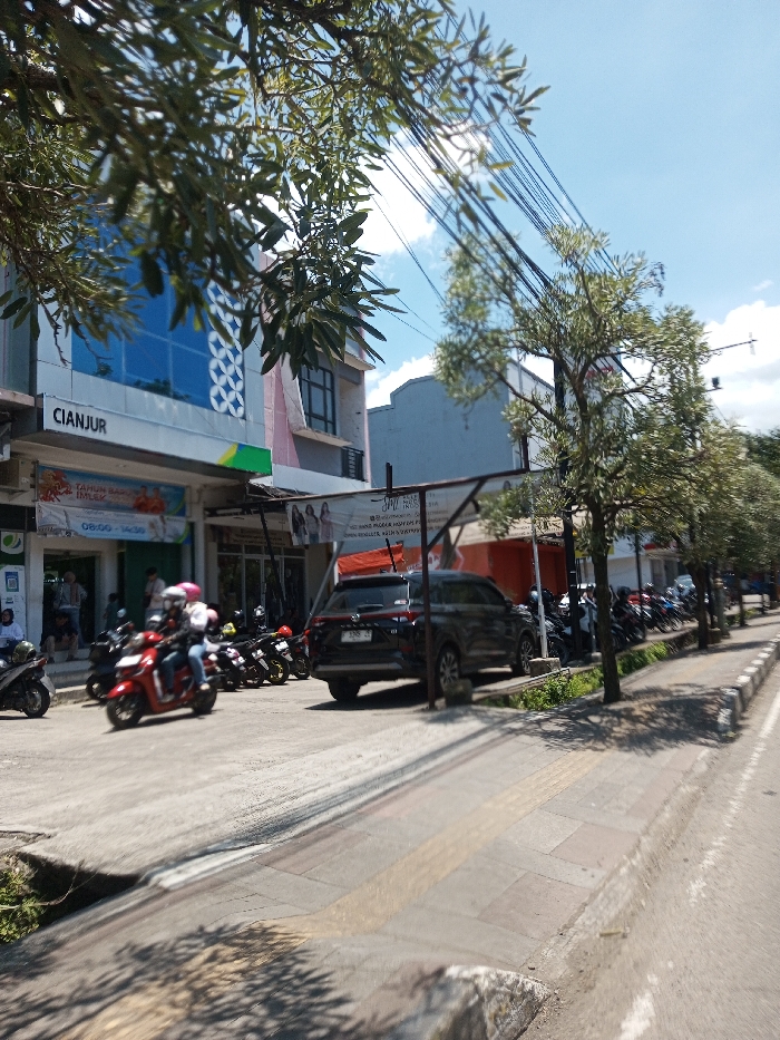 The scene depicts a sunny daytime street view in the city of Tagtse Tashidingkha, Bhutan. In the foreground, a paved sidewalk with yellow tactile paving is visible on the right, bordered by a concrete curb. A red scooter, ridden by two individuals wearing helmets (one white, one pink), moves from left to right on the asphalt road, appearing slightly blurred due to motion. Numerous other motorcycles are parked along the left side of the road and sidewalk. A dark-colored SUV is parked facing away from the viewer, partially beneath a metal awning structure. In the mid-ground, a row of trees with light green foliage lines the right side of the sidewalk, casting shadows. Overhead, multiple black utility lines crisscross a clear blue sky with scattered white clouds. To the left, a multi-story building features blue-tinted windows with white decorative patterns and a fascia bearing the word "CIANJUR." A banner below advertises "Tahun Baru Imlek" with operating hours "08:00 - 14:30." Other commercial signs and parts of additional buildings are visible in the background. Branches with green leaves partially obscure the upper left of the frame. Several indistinct figures are visible near the building entrance.