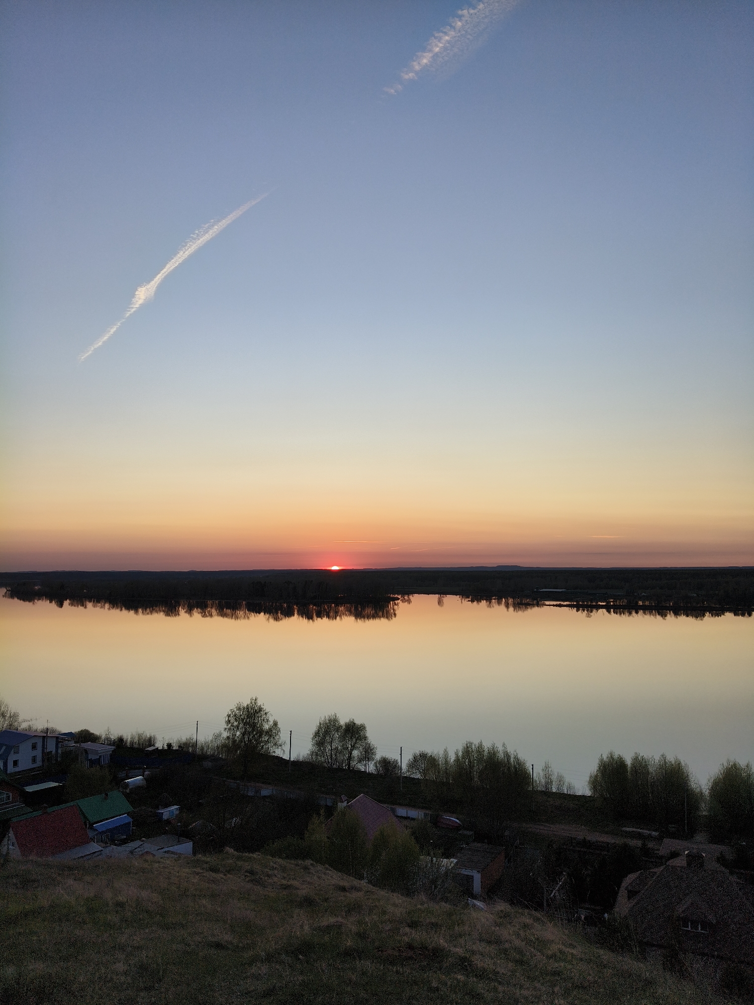 The image shows a landscape view during sunset in Ольховка, Russia. In the foreground, there is a grassy hill, with houses and trees scattered across it. In the middle ground, a large body of water reflects the colors of the sunset sky. The sun is setting on the horizon, creating a warm orange and red glow. Above the horizon, the sky transitions from orange to light blue. There are two contrails in the sky.