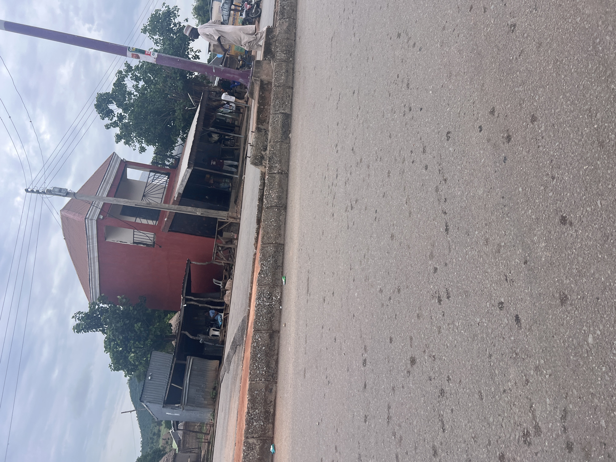 The image depicts a street scene in Kona Garu, Nigeria. The perspective is from ground level, looking outwards towards the street and the buildings lining it. A prominent red building with a white-trimmed roof is visible on the left, featuring an open-air section with a balcony. Adjacent to it, several makeshift stalls with corrugated metal roofs line the street, offering various goods.
Several people are present, including a man in a light-colored traditional outfit standing near a purple pole. Other individuals are seen near the stalls, some appearing to be vendors and customers. A few motorcycles are parked in the background, suggesting local transportation. The ground is asphalt, with some debris and small puddles scattered across its surface. The sky is overcast with grey clouds, indicating a potentially cloudy or rainy day. The overall atmosphere appears to be that of a bustling local market or commercial area. There are no discernable emotions conveyed by the people in the image. Some text is visible on posters on the purple pole, but it is too small and blurry to read.