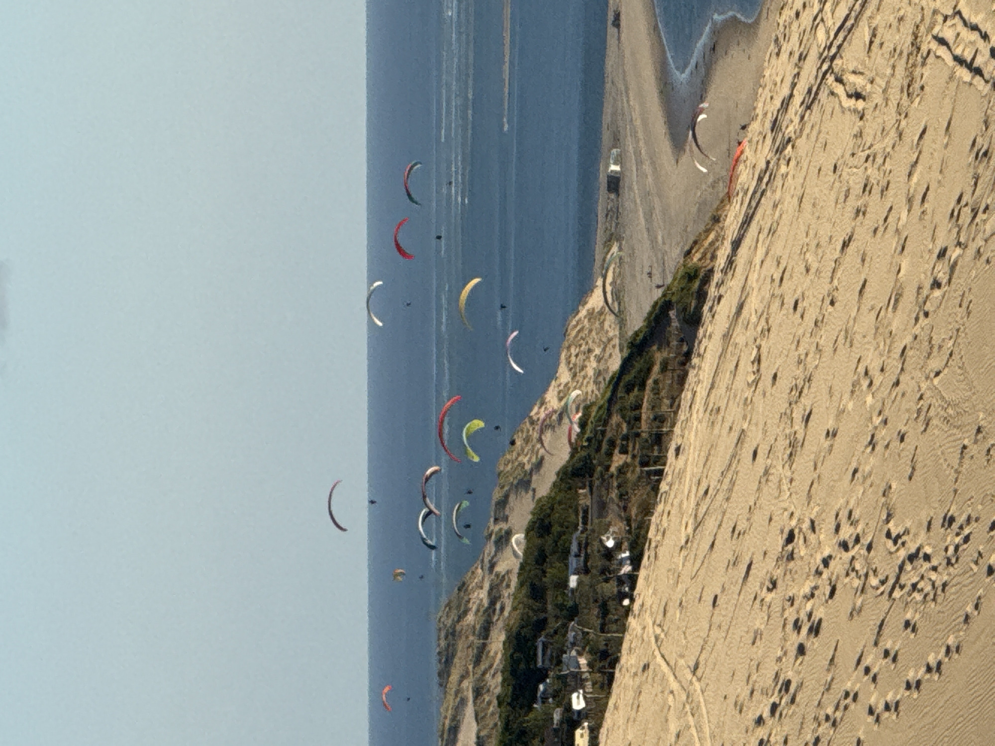 In the city of La Teste-de-Buch, France, a sandy dune slopes down towards a body of water, with a beach and some vegetation visible in the middle ground. The sky is a clear, pale blue. Several paragliders, appearing as colorful canopies with people suspended beneath them, are in various stages of flight. Some are over the water, while others are closer to the dune and the beach. The sand on the dune is marked with footprints and tire tracks, indicating activity. Several small structures, possibly caravans or mobile homes, are situated on the vegetated area near the dune. The overall scene captures a recreational activity taking place in a coastal environment.