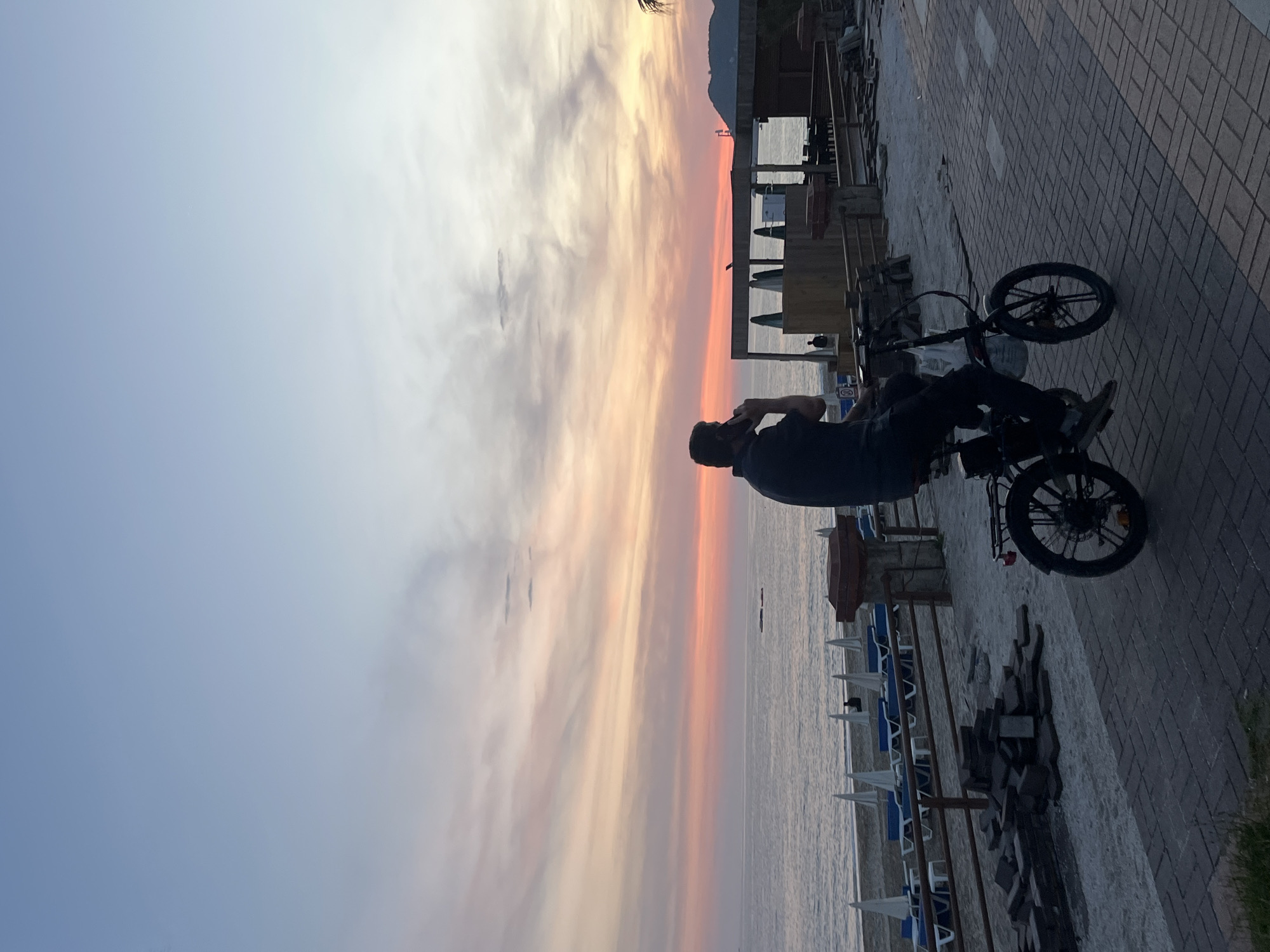 The image shows a man on a bicycle, set against a vivid sunset over the Mediterranean Sea. The scene is likely in Alanya, Turkey, given the coastal location and likely latitude. The sky displays a gradient of colors from pale blue to vibrant orange and pink near the horizon. The man is seen sitting on his bicycle, which is parked on a paved walkway next to the beach. Rows of beach chairs with white and blue accents are visible along the shore. In the background, there are building structures with open areas, possibly part of a beachfront establishment. The overall lighting suggests the photo was taken during the golden hour, shortly after sunrise or before sunset.