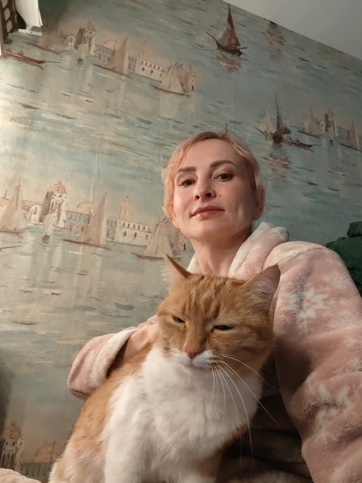A woman with short, blonde hair is holding an orange and white cat. She is wearing a pink robe with a white snowflake pattern. The cat is looking down and to the left, with its eyes partially closed. The woman is looking towards the camera with a slight smile. Behind them is a wall with a mural depicting a harbor scene with sailboats and buildings. The mural has a muted color palette of blues, greens, and tans. The lighting in the image is soft and natural. The scene appears to be indoors. The location is not specified in the image or provided context.