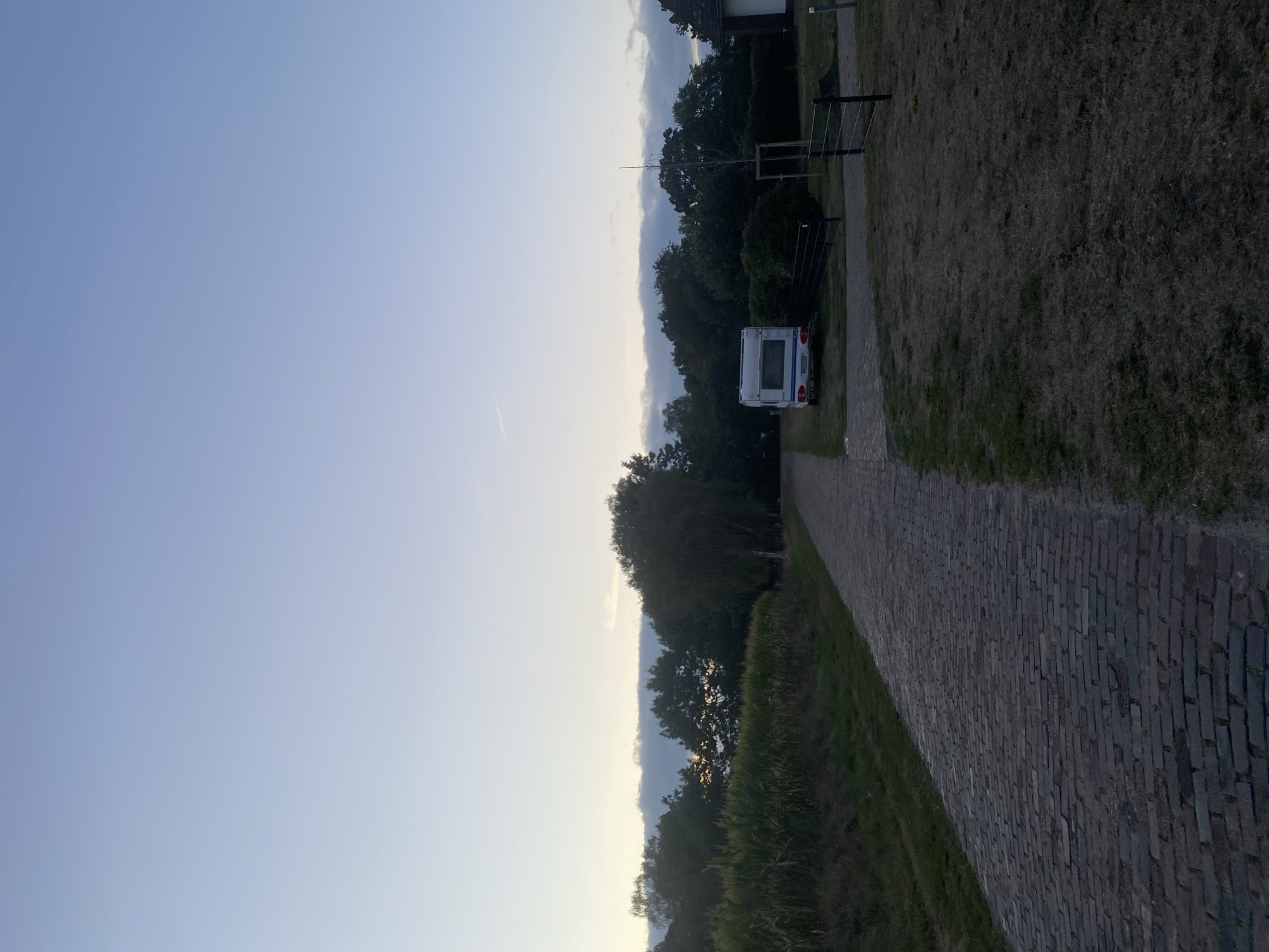 The image depicts a rural scene at dusk in Schwarme, Germany. A cobblestone path leads into the distance, flanked by a field of tall cornstalks on the left and dry grass on the right. In the middle ground, a white caravan is parked on a paved area adjacent to the path. Beyond the caravan, a line of trees forms a dark silhouette against the pale sky. A dark fence runs along the right side of the caravan. The sky is a gradient of light blue, fading to a lighter hue near the horizon, with a few wispy clouds. The overall impression is one of tranquility and early evening in a countryside setting.