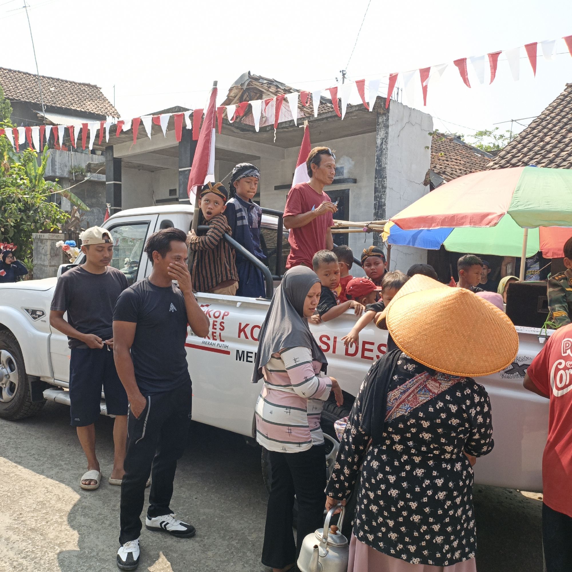 In Tanahbaya, Indonesia, on a sunny day, a group of people are gathered around a white pickup truck, likely for a local celebration or event. Red and white pennant flags are strung across the scene, adorning the roofs of houses and the truck itself. Several children are positioned in the bed of the truck, some wearing traditional headwear. Two adult men stand near the front of the truck, one wearing a black t-shirt and the other a dark grey t-shirt and a beige cap. A woman in a grey hijab and a striped long-sleeved shirt is walking beside the truck, carrying a metal teapot. Another woman, wearing a black floral dress and a wide-brimmed straw hat, is seen from behind, also near the truck. The truck has text printed on its side, with some letters visible including "KOP," "ES," "KEL," "KO," "SI," "DES," and "MERAH PUTIH." The ground is paved and shows evidence of shadows. The background consists of houses with tiled roofs and trees.