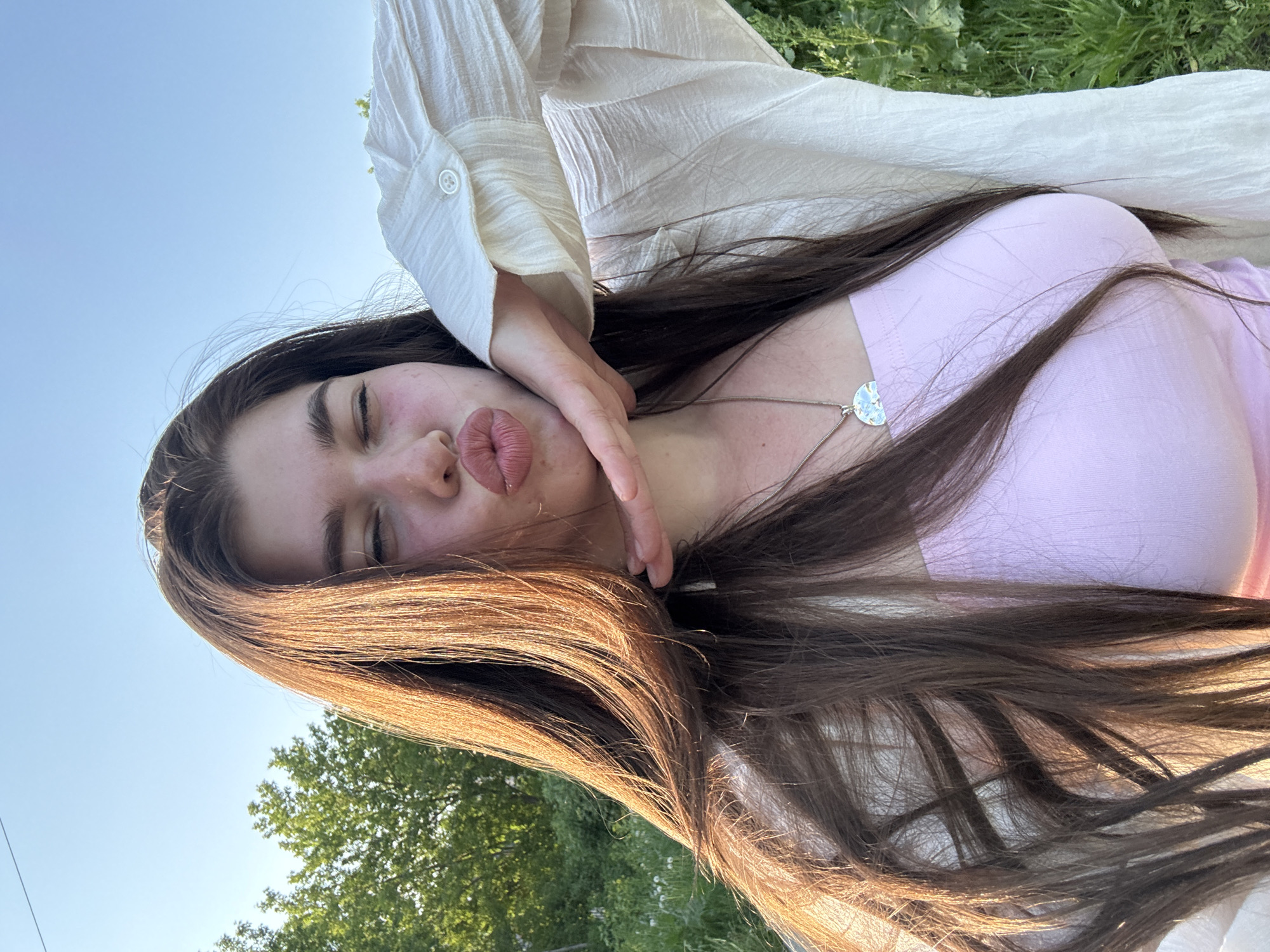 A young woman is photographed outdoors in natural sunlight, likely in Troshkovo, Russia. She has long, dark brown hair, a fair complexion, and is wearing a light pink top and a white button-down shirt. She's making a "duck face" expression with her lips. Her left hand is near her face. A silver necklace is visible around her neck. The background features green vegetation and a clear blue sky.