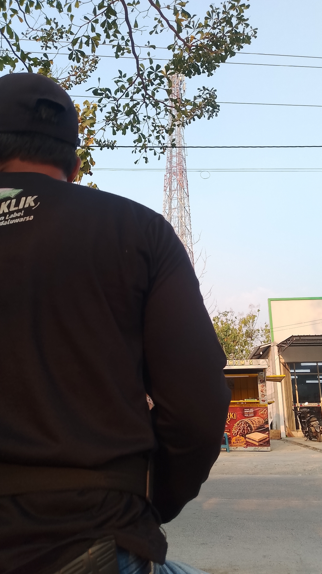 In Karanglangu, Indonesia, a person wearing a black long-sleeved shirt and a black baseball cap is standing outside. The person is facing away from the camera, and their back is partially visible. In the background, a tall transmission tower stands against a clear blue sky. A tree with green leaves is positioned in the foreground, partially obscuring the view of the tower. To the right of the tower, a small food stall with yellow and white signage is visible, and a motorcycle is parked in front of a building. The ground in front of the stall and building appears to be paved. The overall scene suggests an outdoor setting in a populated area, possibly a street or a market.