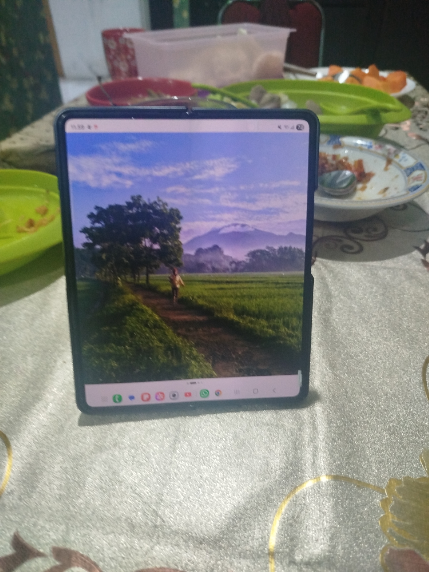 The image shows a smartphone, folded in a book-like manner, resting on a textured, light-colored tablecloth. The smartphone screen displays a vibrant landscape wallpaper, featuring a dirt path leading through lush green rice paddies towards a cluster of trees and distant mountains shrouded in a light haze under a clear blue sky with scattered clouds. On the top bar of the screen, the time "11:38" is visible, along with battery and signal indicators. The bottom of the screen shows a row of application icons.
Surrounding the smartphone, on the same tablecloth, are elements of a meal. To the left, a bright green plastic bowl contains food. Further back and to the right, a partially eaten meal is visible on a white plate with a blue and gold decorative rim. Another green plastic serving dish holds what appear to be pieces of fruit, possibly papaya, and some meatballs. In the background, a red and white patterned cup is visible, along with the edge of a translucent plastic container. The overall scene suggests a casual dining setting, possibly a home or a modest eatery, with the smartphone positioned centrally as a focal point. The ambient lighting suggests it could be daytime.
The scene is set on a tablecloth that has a textured, almost shimmering surface, with some gold-colored decorative patterns visible in the lower right portion of the image. The lighting appears to be natural, casting soft shadows and highlighting the textures of the tablecloth and the objects on it. There are no people clearly visible in the image, but the presence of a meal implies recent or ongoing activity. The image is taken from a slightly elevated angle, looking down at the smartphone and the surrounding items.
The specific location is a home or restaurant in Bandungrejo, Indonesia.