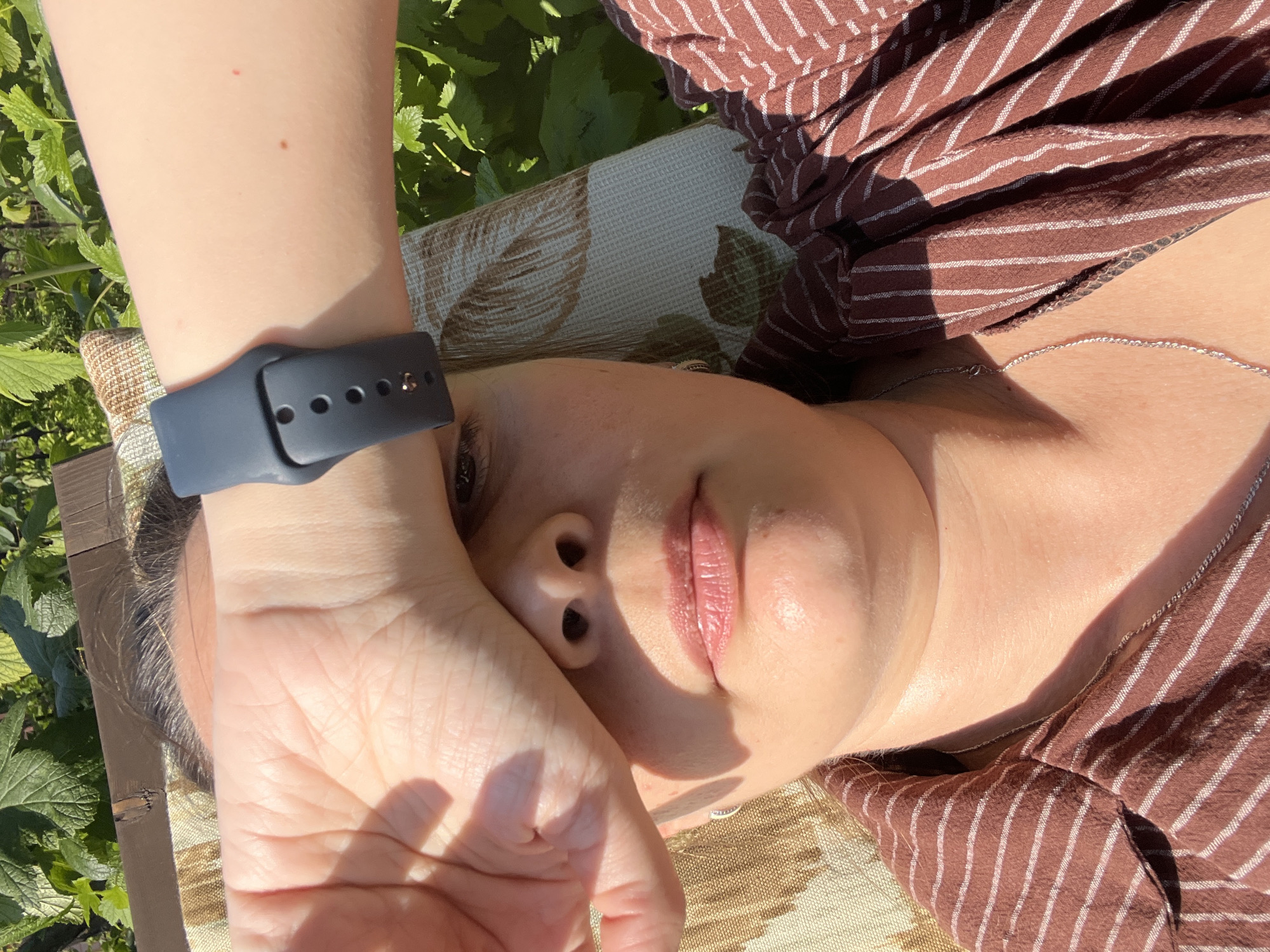 A close-up, overhead shot shows a woman shielding her eyes from the sun with her right hand. She is lying down, possibly on a lounge chair, indicated by the textured fabric visible in the background. Her left arm, adorned with a dark smartwatch, is also raised, partially obscuring her face. Her lips are slightly parted and appear to have lip balm applied. She is wearing a brown striped shirt with white pinstripes and a silver necklace. The environment appears to be outdoors, with green foliage visible behind her, suggesting a garden or natural setting. The lighting indicates it is daytime and sunny. No other individuals are present. The image was taken in Lopatino, Russia.