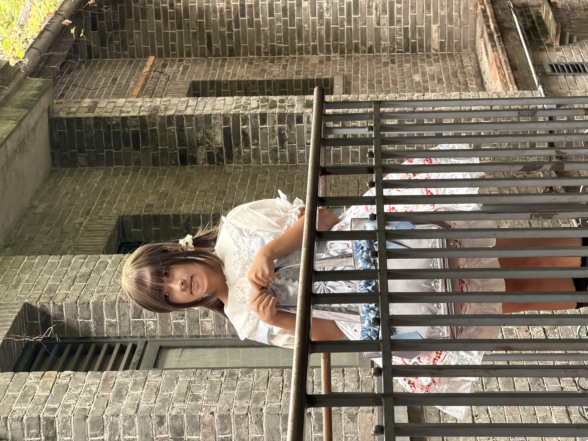 A young East Asian woman with brown hair styled in pigtails, adorned with white flowers, stands on a balcony behind a gray metal railing. She is wearing a white dress with a ruffled collar and puffed sleeves, and a light blue Lolita-style dress with white ruffled trim and red accents. She holds a light blue handbag with white and red details. The background consists of a brick building with a textured gray brick facade and dark windows. The setting appears to be in Chancheng District, China.