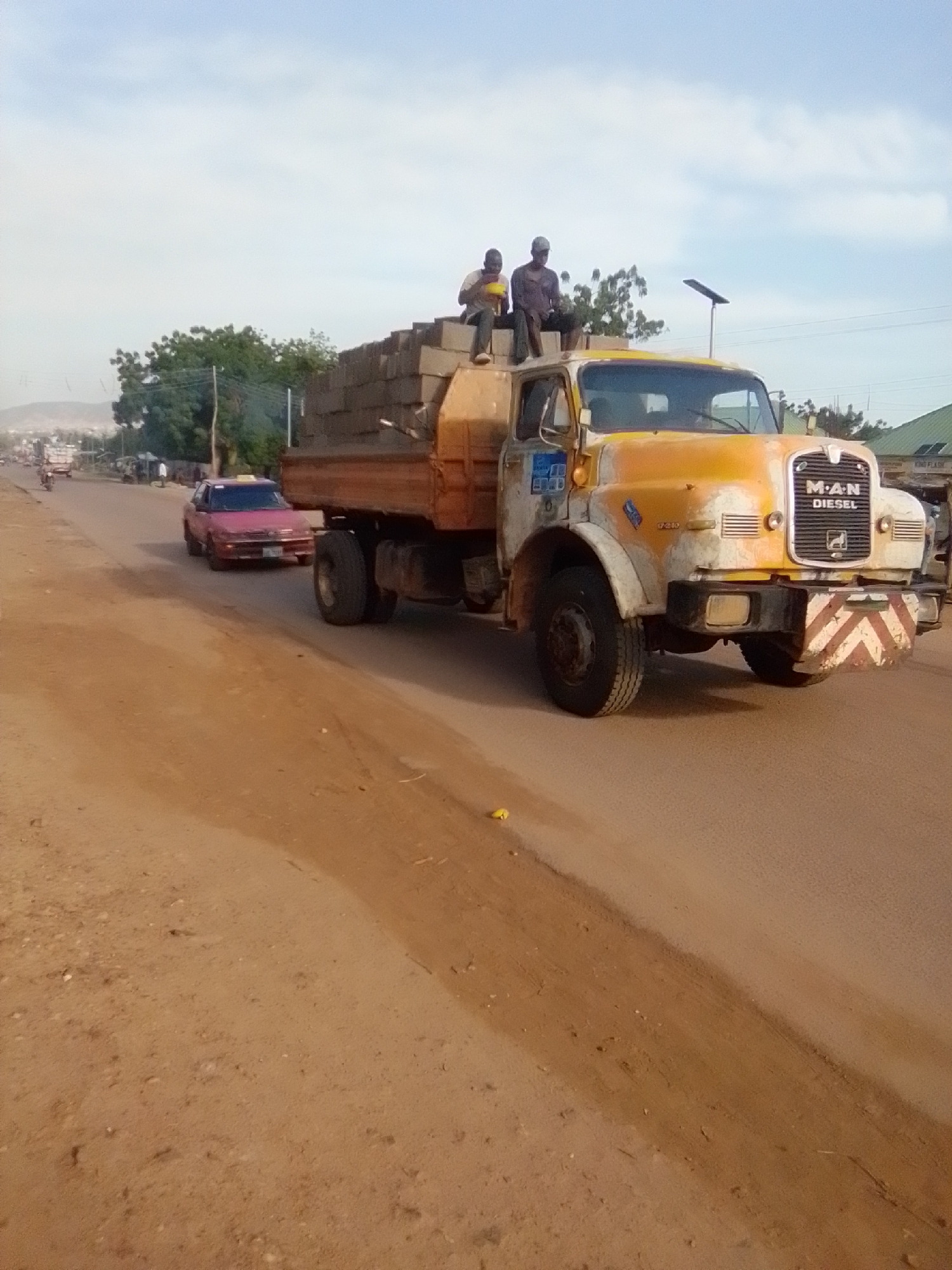 In Galadima, Nigeria, a large yellow and white MAN diesel truck is parked on a dirt road, its flatbed loaded with what appears to be construction bricks. Two men are seated on top of the bricks in the truck bed. One man is wearing a dark shirt and a light-colored cap, while the other is in a light-colored shirt and a dark cap. A red car is driving past the truck on the left side of the road. The sky is partly cloudy. In the background, there are trees and buildings. The overall scene depicts a typical day in a Nigerian town, with commercial activity and transportation.