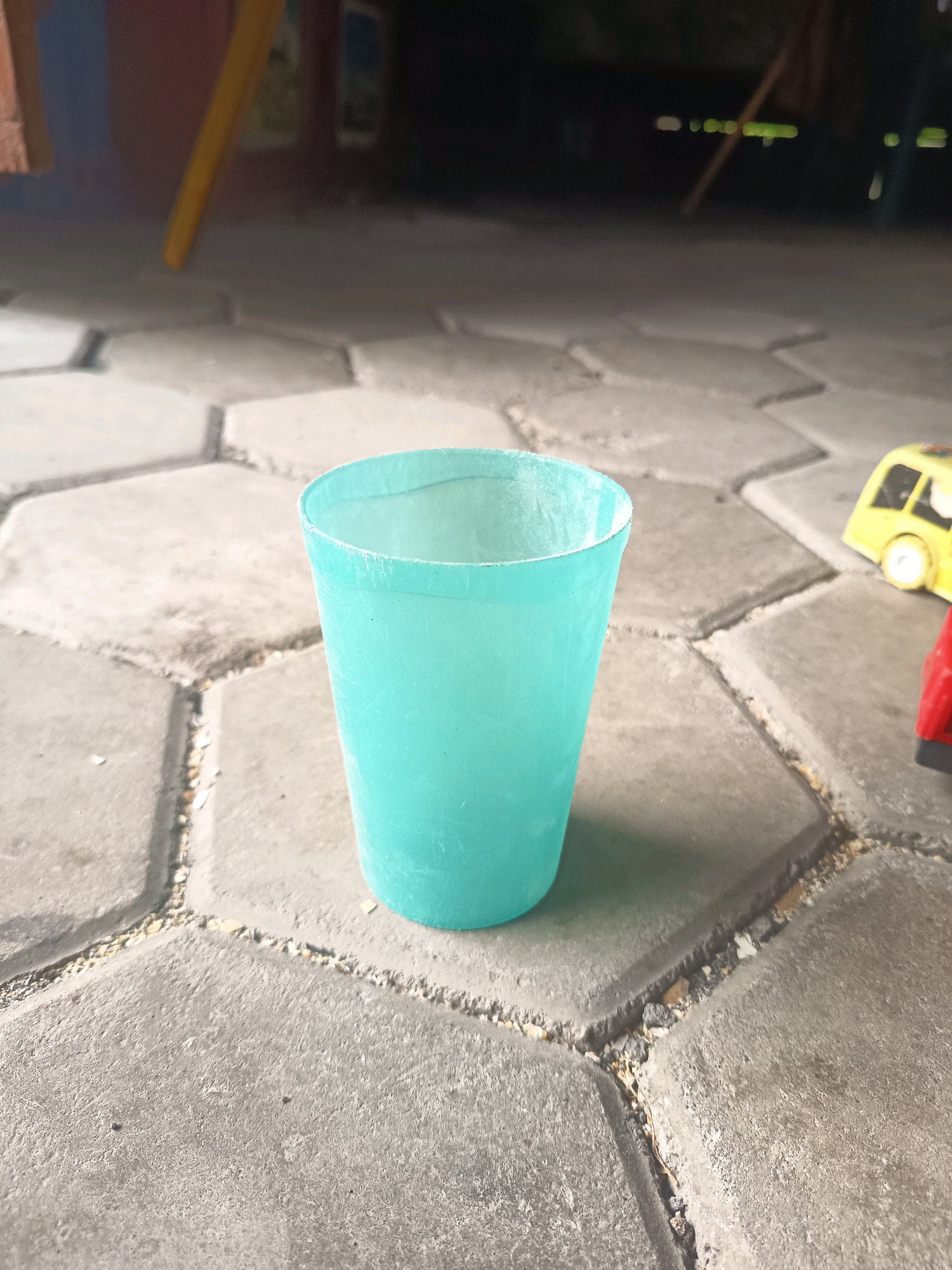 A turquoise plastic cup sits on a patterned concrete surface in what appears to be an outdoor area in Sidorejo, Indonesia. The concrete is composed of interlocking hexagonal and rectangular pavers. In the background, a blurred yellow toy car and a red object, possibly another toy, are visible. The lighting suggests it is daytime, with some ambient light reflecting on the cup and the paved surface. The environment appears to be an outdoor space, possibly a yard or a public area, given the paved ground and the presence of children's toys.