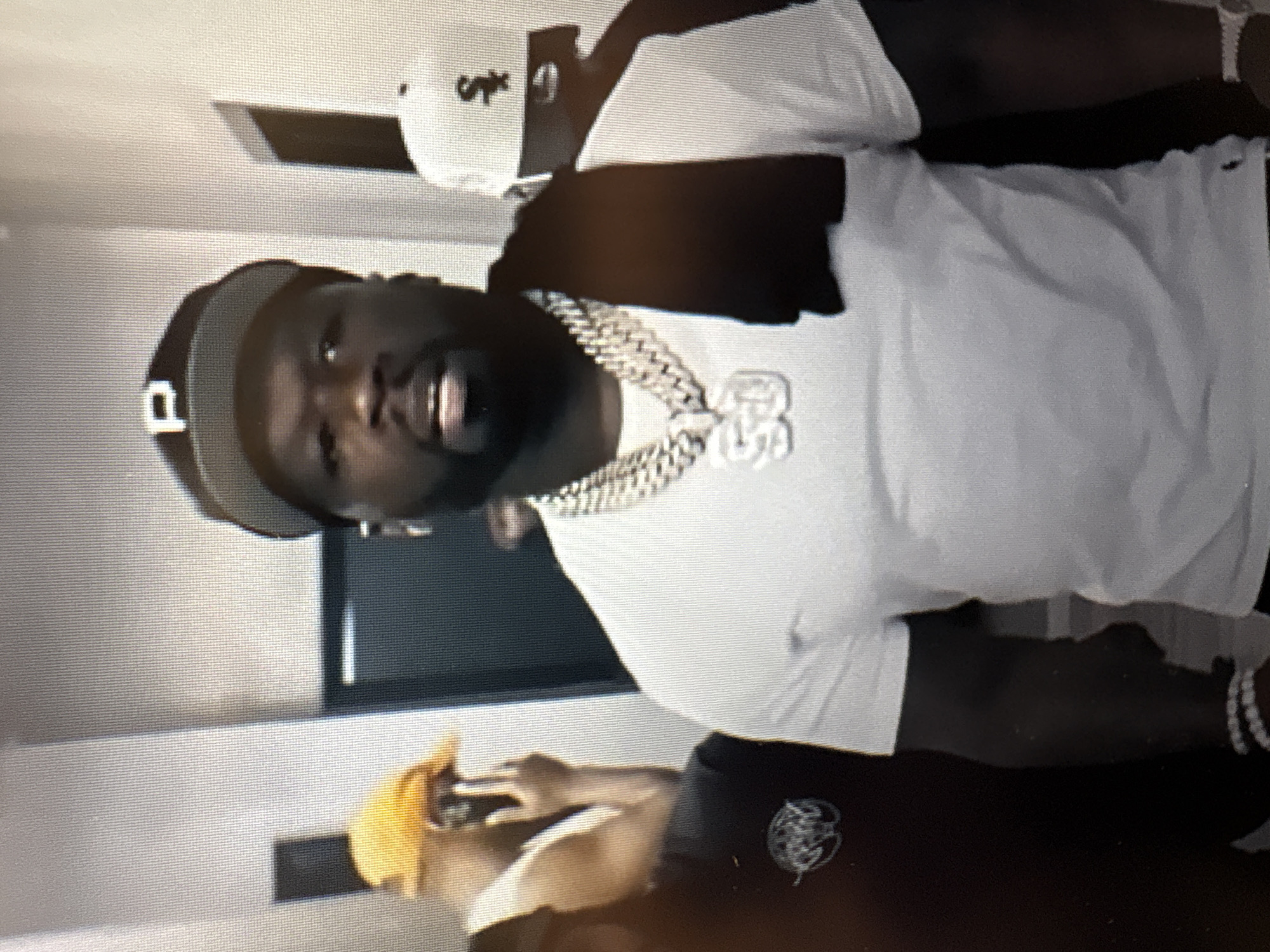 The image shows a man wearing a white t-shirt, a black baseball cap with a white "P" on it, and a large diamond chain with a pendant that appears to say "50". He is also wearing a black cloth draped over his shoulders. The man is standing indoors in what appears to be a hallway or corridor with light-colored walls. There are other people partially visible in the background, including one wearing a yellow cap and another wearing a white baseball cap with a black logo. The image is a close-up shot focused on the man in the center. There is no indication of the location being Sofia, Bulgaria.