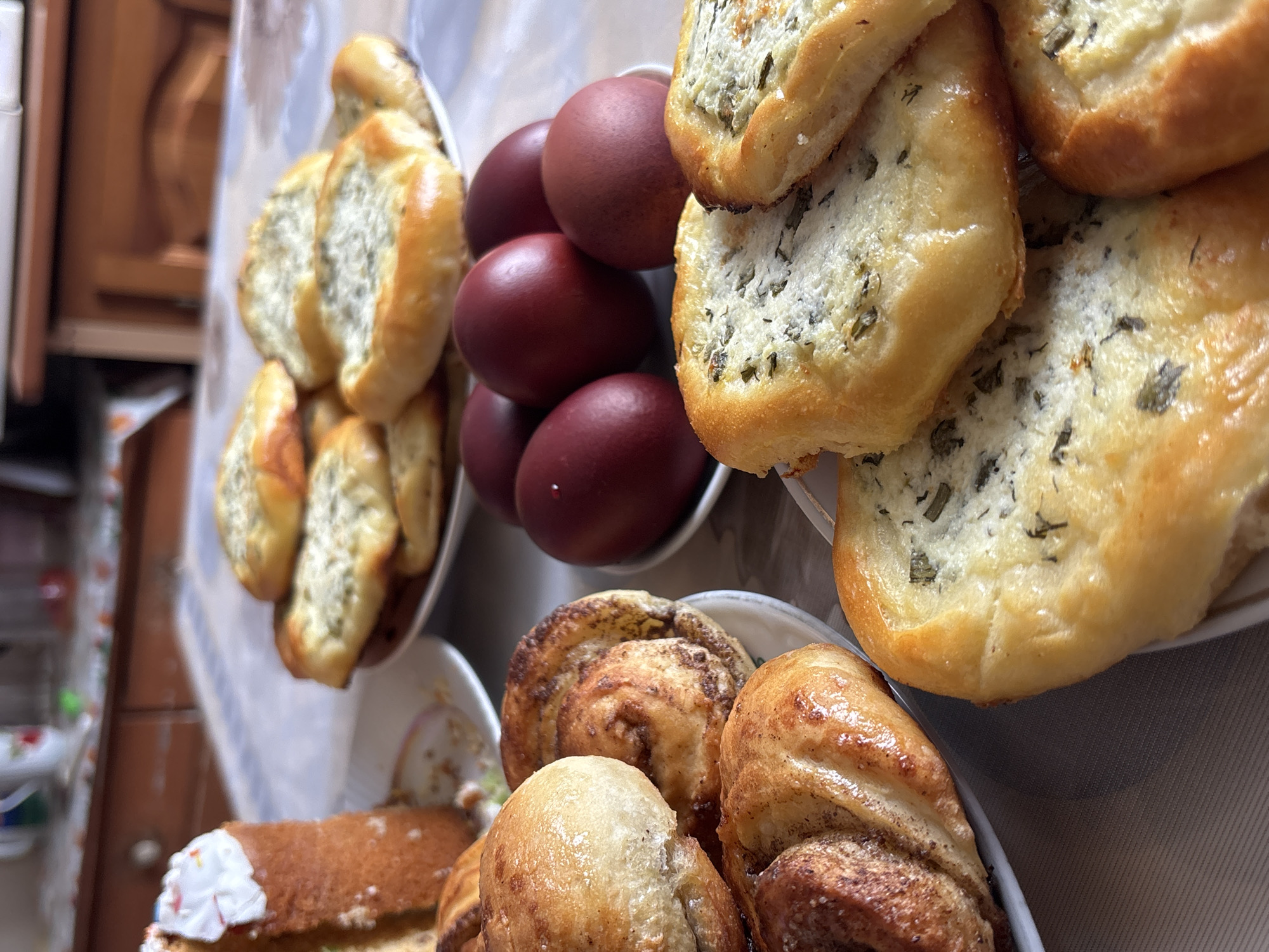 The image shows an assortment of baked goods and dyed eggs on a table. The food items include several plates of buns, some with a savory filling featuring green herbs, and others that appear to be cinnamon rolls. A slice of decorated cake is also visible in the bottom left. A small bowl contains dark red, possibly Easter eggs. The environment appears to be indoors, perhaps a home setting with wooden cabinetry visible in the background. The image is taken at a close range, and the focus is primarily on the food items.
There is no information to suggest that the image was taken in Urada, Russia, so this location is not included in the description.
