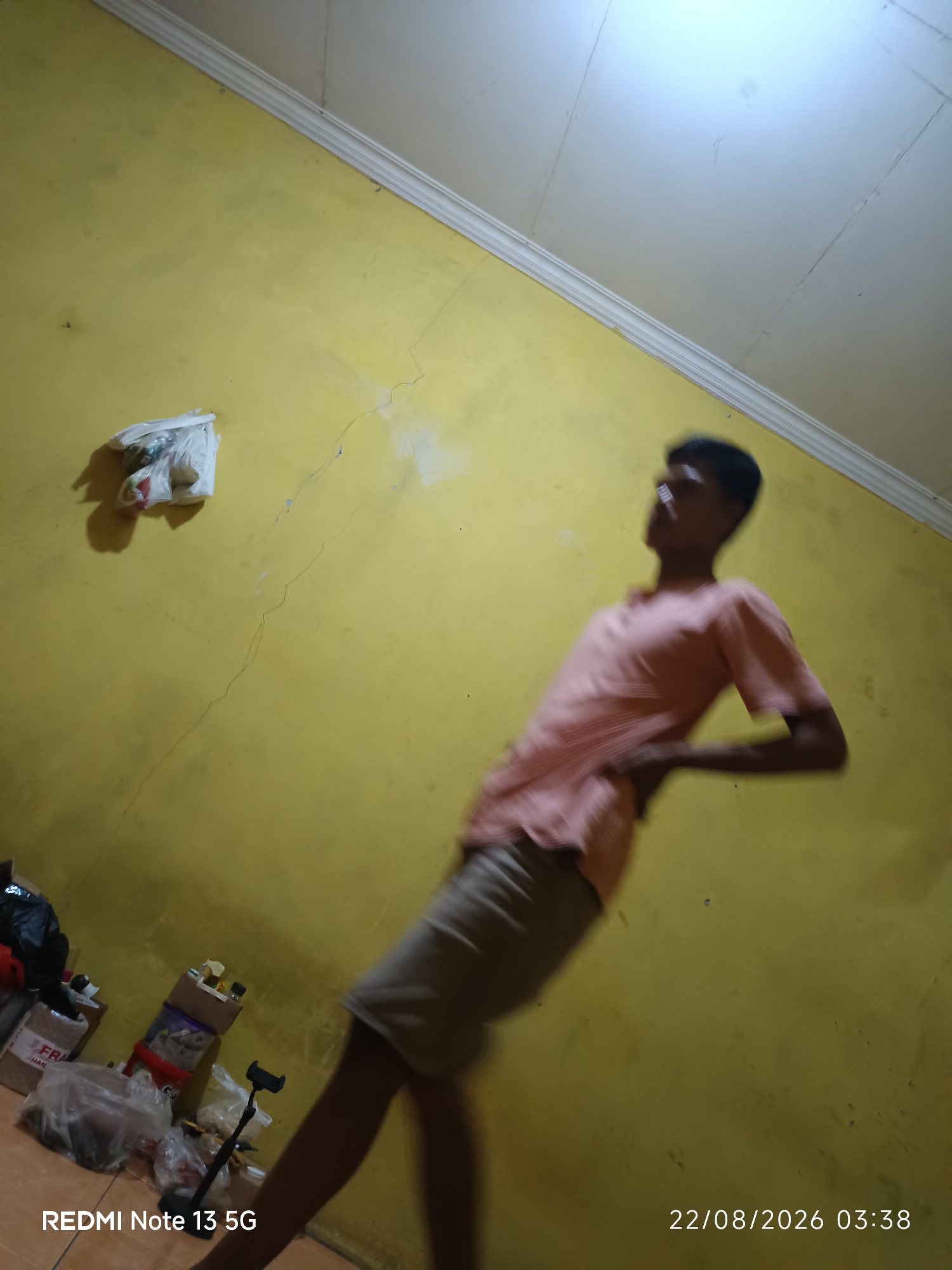 The image displays an indoor scene with a yellow-painted wall and a white ceiling. A person, appearing to be a young male, is captured in motion, leaning backward and to the side, wearing a light pink polo shirt and beige shorts. The image also shows a plastic bag hanging on the wall, several boxes and bags of items on the floor, and a cell phone tripod. A timestamp in the lower right corner indicates "22/08/2026 03:38" and the text "REDMI Note 13 5G" in the lower left corner. The overall impression is of a candid, possibly staged, snapshot taken in a residential or storage area. The location is not explicitly stated within the image itself, so its placement in Talaga Sari, Indonesia, cannot be confirmed from the visual information provided.