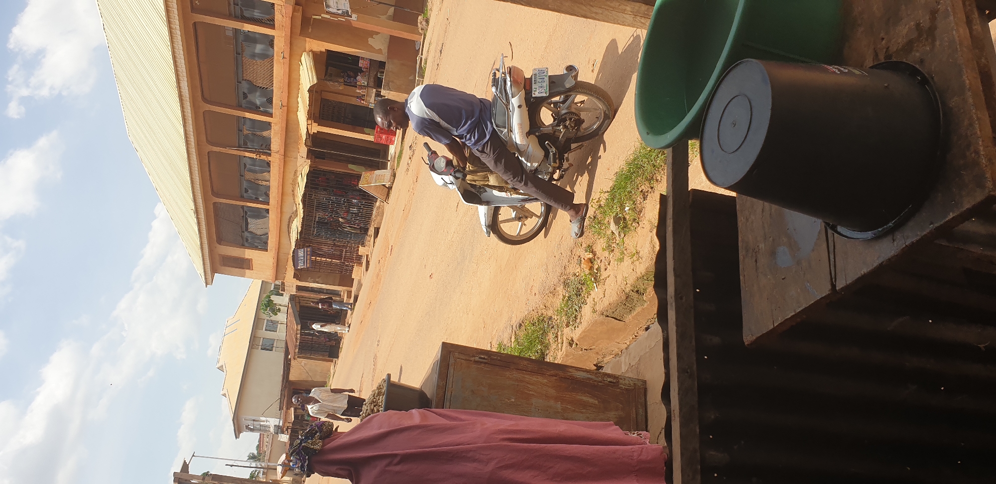 In the city of Sabon Tasha, Nigeria, a man rides a white motorcycle on a dirt road lined with buildings. The man, wearing a dark blue shirt with white accents and dark pants, is seated on the motorcycle, looking to his left. The motorcycle has a license plate that reads "RSH-641QF". In the foreground, to the left, a woman wearing a pink headscarf and dress is partially visible, standing next to a metal cabinet filled with produce. A large dark bucket is positioned on a wooden surface in the lower right of the image, next to a large green basin. The buildings in the background are two stories with orange facades and metal-grated windows. The sky is partly cloudy.
