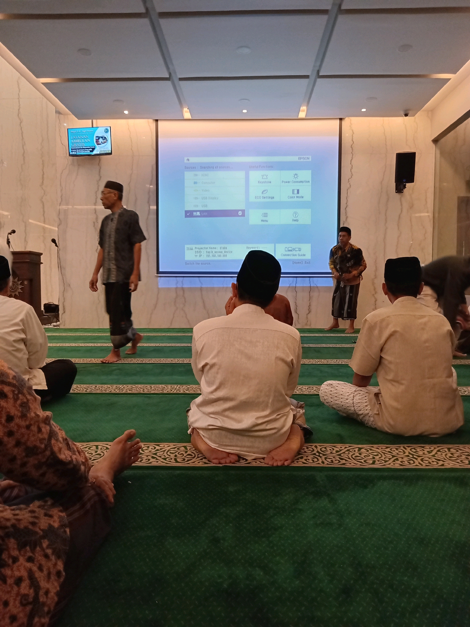 In Randuagung, Indonesia, an indoor scene captures individuals seated on a green carpet, presumably within a mosque or a prayer hall, facing a large projector screen displaying an Epson menu with options like "Keystone," "Power Consumption," "ECO Settings," and "Color Mode." Several men, some wearing traditional Indonesian attire and caps, are visible. One man is walking across the carpet in the middle ground, while others are seated in the foreground and background, facing away from the camera. The room features white marble-patterned walls and a white ceiling with recessed lighting. A smaller screen showing text and an image is mounted on the left wall. The overall atmosphere appears to be a presentation or lecture, given the presence of the projector screen and the seated audience.