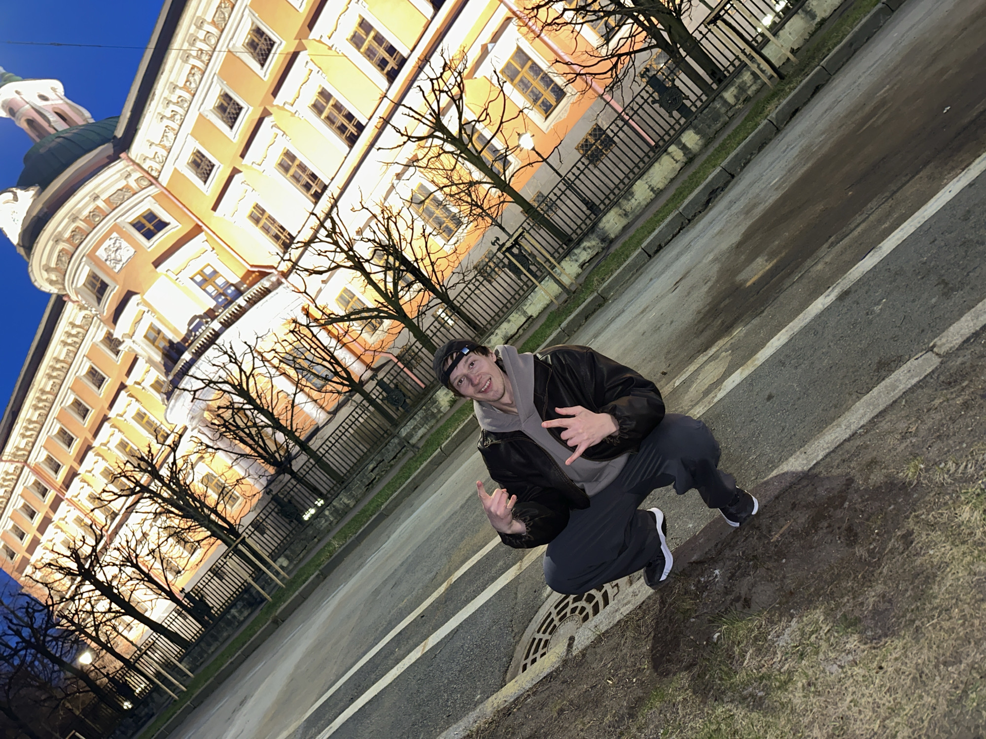 A young man is crouching next to a roadside drain, making hand gestures. He is wearing a black jacket, gray hoodie, black sweatpants, and black and white sneakers. The backdrop is a large, multi-story building with yellow and white facade, multiple windows, and a domed tower on the left. Bare trees line the street in front of the building, partially obscuring it. A black metal fence runs along the edge of the sidewalk. The location is the Smolny Cathedral in Saint Petersburg, Russia. The lighting suggests it is nighttime.