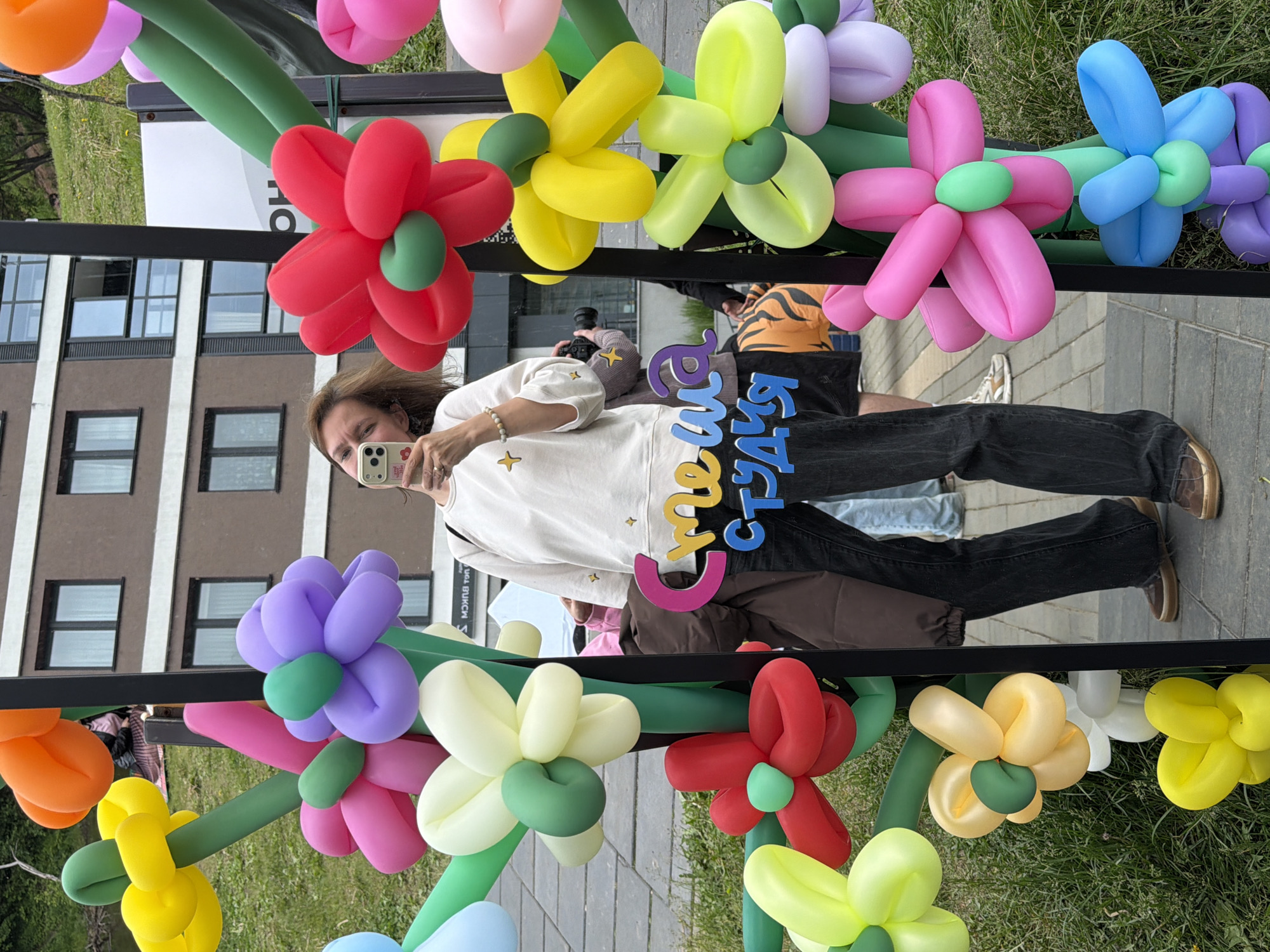 The image captures a woman taking a mirror selfie outdoors in Izhevsk, Russia, framed by an archway of colorful balloon flowers. The woman, with light brown hair, is positioned centrally within a large, rectangular mirror with a thin black frame. She wears a cream-colored long-sleeved sweatshirt adorned with small gold star appliques, dark wide-leg jeans, and brown lace-up shoes. She holds a white iPhone, likely a Pro model with multiple cameras, in her right hand, angled to capture her reflection in the mirror. A light-colored beaded bracelet is visible on her left wrist.
The mirror is surrounded by an elaborate arrangement of balloon flowers, constructed from twisted balloons, creating a vibrant, multi-colored border. These flowers feature various colors including red, orange, yellow, lime green, dark green (used for stems and centers), pink, purple, light blue, and white/cream. Some balloon flowers are rooted in the grassy foreground, while others appear to be part of a freestanding structure.
In the background, reflected in the mirror, are modern multi-story residential buildings with brown and white facades and numerous rectangular windows. A grassy slope or hill is visible beyond the buildings on the left. To the right of the woman's reflection, a white vertical sign with black Cyrillic text, partially legible as "МОН," is visible. Below this, a blue and yellow sign reading "Смеша СТУДИЯ" (Smesha Studio) in Cyrillic script is reflected across the woman's lower torso.
Reflected behind the primary woman, another individual is seen holding a black camera with a lens, also seemingly taking photos. This person wears a grey long-sleeved top with gold star appliques, similar to the primary woman's. Parts of other individuals, including legs and shoes, are faintly visible in the deeper reflections. A striped object, possibly a tiger-patterned bag, lies on the ground to the right of the woman's reflected legs. The ground consists of grey interlocking pavers, with patches of green grass surrounding the balloon flower display. The scene suggests an outdoor public event or a decorative installation.