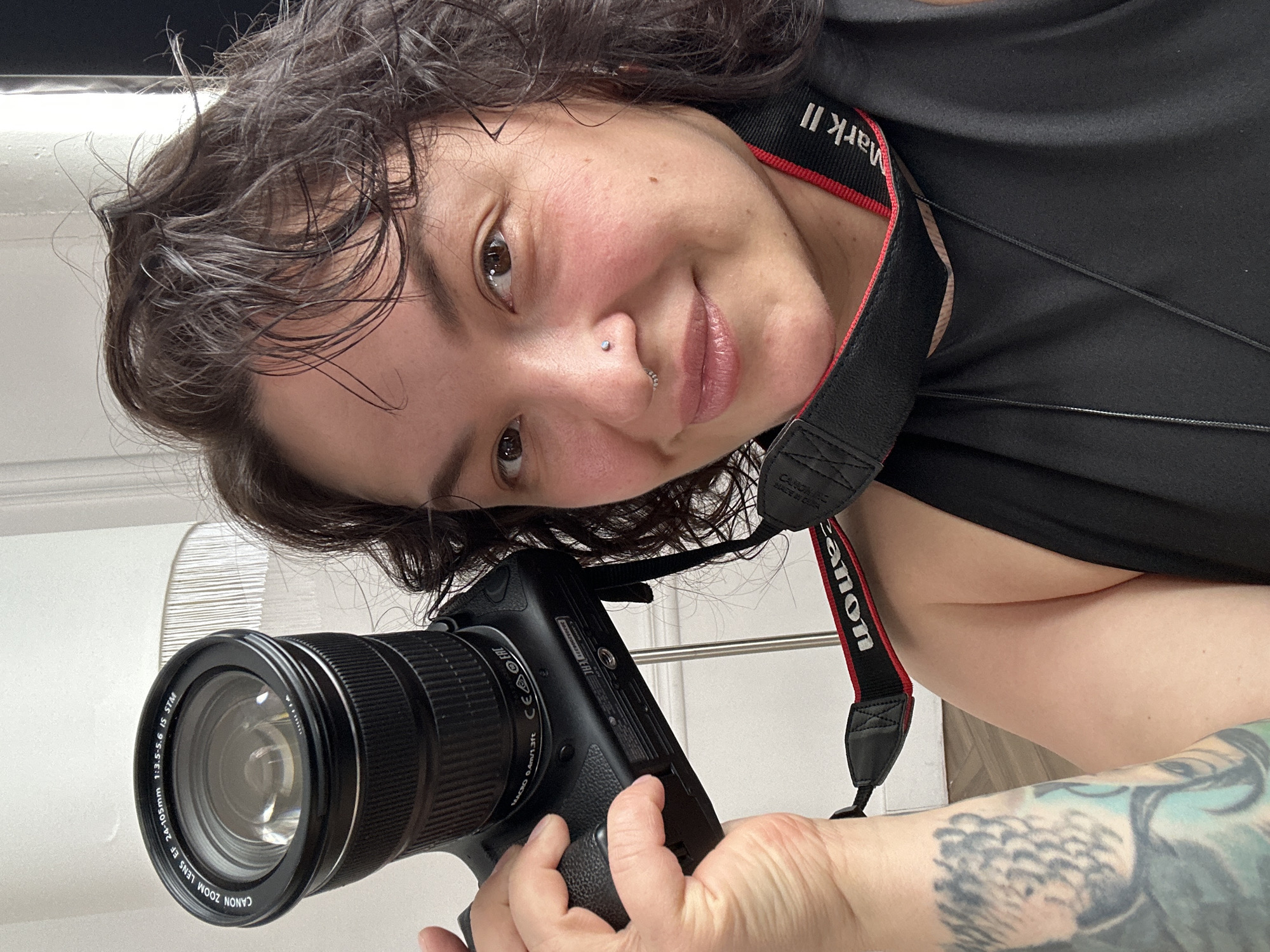 A woman, located in Chaikovsky, Russia, is captured in a close-up, angled selfie, smiling gently. She has dark, wavy hair, dark eyes, and fair skin with a slight flush on her cheeks. She wears a small stud piercing on her nose bridge and a ring on her right nostril. Her left arm, which is visible, is adorned with tattoos. She is wearing a black top and holds a black Canon DSLR camera, angled as if she is taking the photo herself.
The camera's lens is a "CANON ZOOM LENS EF 24-105mm 1:3.5-5.6 IS STM" with a "MACRO 0.4m/1.3ft" marking. The camera body displays "CE FC" certification marks and the number "303052008". A red and black camera strap, visible around her neck, is marked with "CANON", "MADE IN CHINA", and "Mark II", suggesting a Canon Mark II series camera. The background is a plain white wall, indicating an indoor setting.