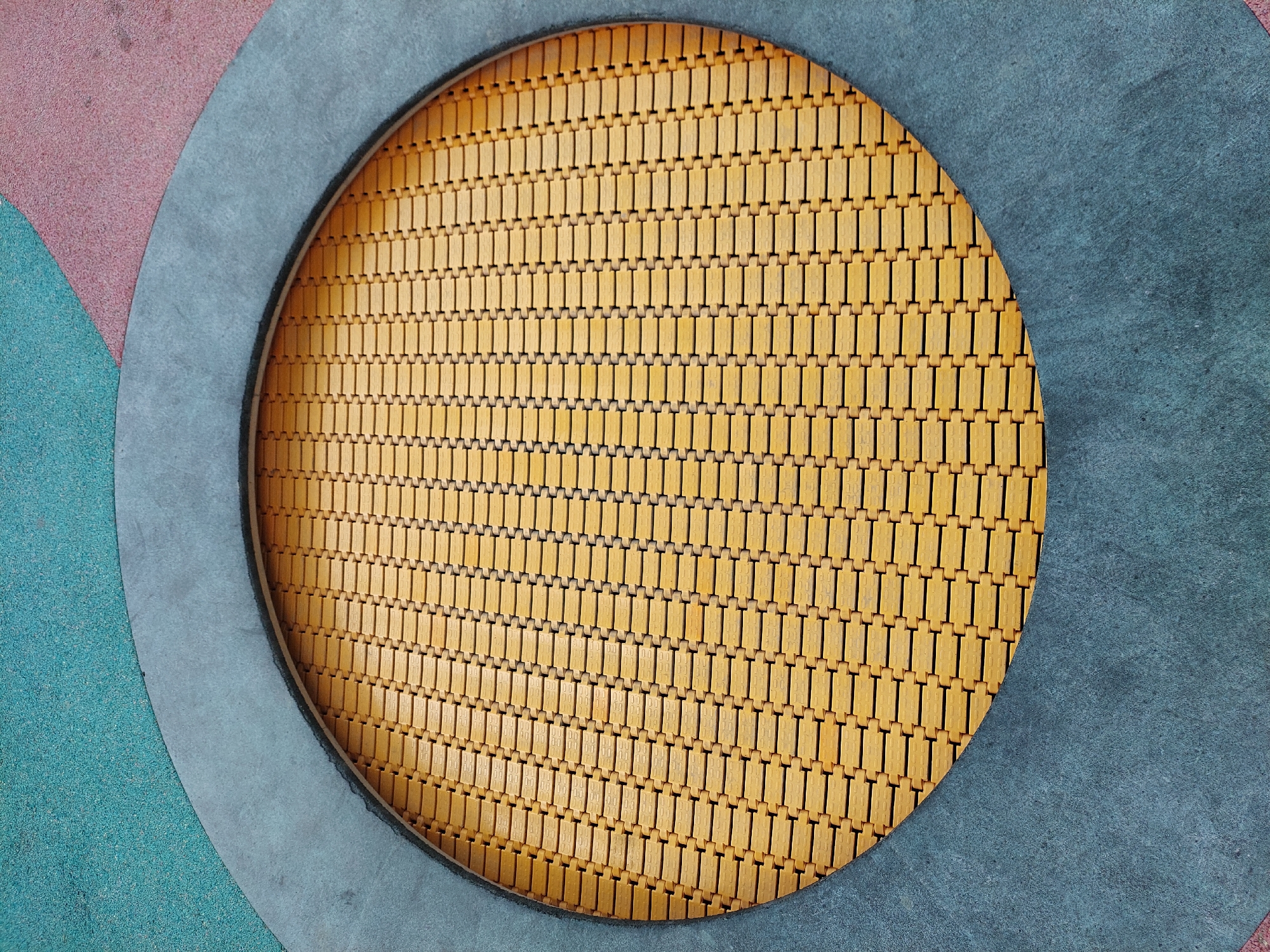 This is a high-angle, close-up shot of a circular trampoline embedded in a playground surface. The trampoline bed is made of interlocking yellow rubber or plastic tiles arranged in a grid pattern. A thick, dark gray border surrounds the trampoline. The playground surface surrounding the gray border is made up of irregularly shaped sections of textured material in teal and pinkish-red. The scene is devoid of people and any other discernible objects. This image likely depicts a public playground area, possibly within the city of Alikonovka, Russia, given the context provided.