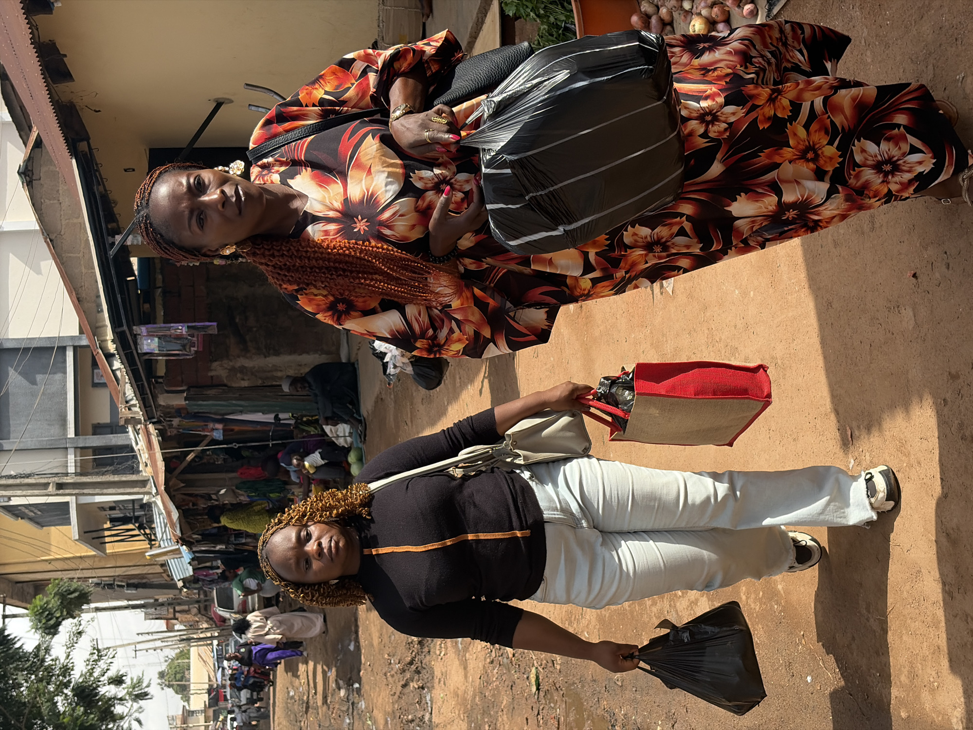 The image shows two women in what appears to be a bustling market in Jos, Nigeria. The photograph is oriented vertically.
The woman in the foreground is wearing a black long-sleeved shirt with an orange stripe across the chest and light-colored pants. She has curly brown hair and is holding a black plastic bag in her right hand. A beige shoulder bag is slung across her body, and a red and beige tote bag is placed on the ground to her right. She is looking towards the camera with a neutral expression.
The woman in the background is wearing a vibrant dress with a floral pattern in shades of orange, black, and white. She has braided hair and is wearing large gold earrings and rings on her fingers. She is holding a large black plastic bag and is also looking towards the camera, with a slight smile.
The scene depicts an outdoor market with stalls visible behind the women, selling various goods. The ground is dusty, suggesting dry weather. The sunlight indicates it is daytime, likely mid-morning or afternoon. The overall atmosphere appears to be lively, typical of a busy marketplace.
