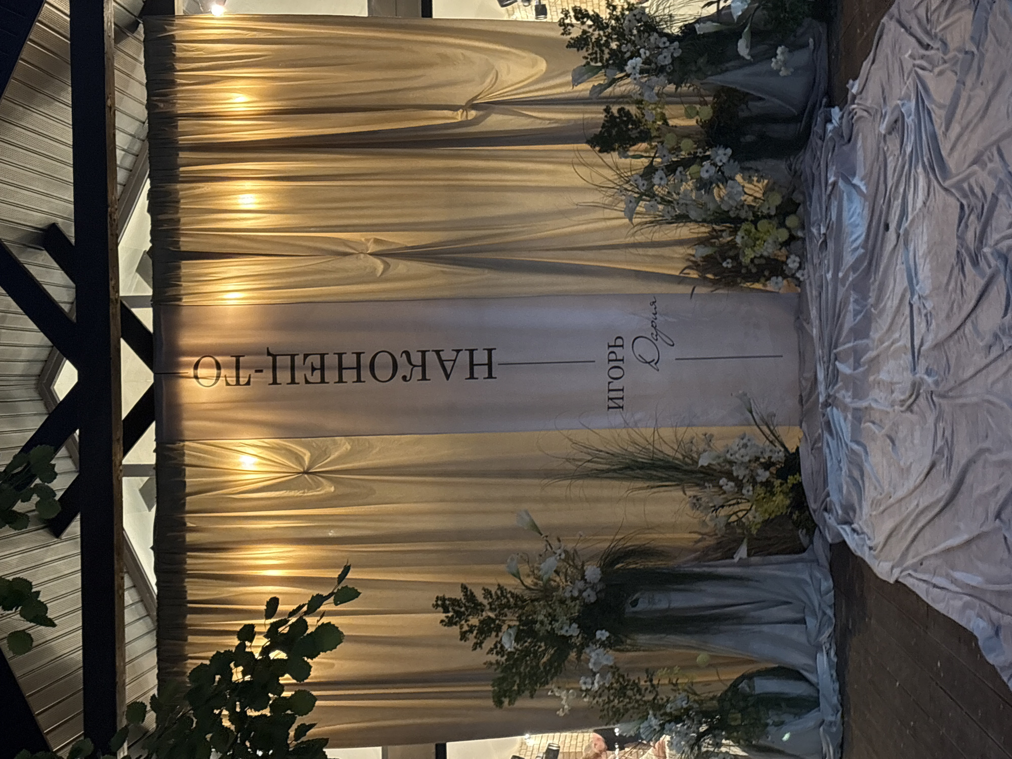 The image captures an event setup, described as being in Perm, Russia, likely an evening or nighttime scene. A covered overhead structure features a white corrugated ceiling and dark beams. Green foliage from a tree or plant is visible in the upper left foreground, partially obscuring the ceiling.
The main subject is a decorative backdrop consisting of light beige-gold curtains, illuminated by warm lights from behind, creating vertical shafts of light. Centered on these curtains is a vertical white banner. The banner displays the Russian text "НАКОНЕЦ-ТО" (FINALLY) at the top in dark capital letters, followed by a horizontal line, and then the name "ИГОРЬ" (IGOR) below it, with "Дария" (Daria) written in a cursive script beneath Igor's name.
On either side of the banner, large floral arrangements are placed on the floor. These arrangements contain white lilies, various white and light yellow flowers, and abundant green foliage, including tall grasses. Dark vases or containers support these arrangements, from which light grey fabric is draped onto the floor.
The floor beneath the setup is dark brown wood. A wide, light grey/silver fabric runner is laid out in the foreground, extending from the base of the floral arrangements towards the viewer. The fabric is rumpled and covers a significant portion of the wooden floor.
A blurry human figure is partially visible on the far left, behind the left floral arrangement, obscured by shadows and the decor. The overall lighting suggests an artificial, warm ambiance.