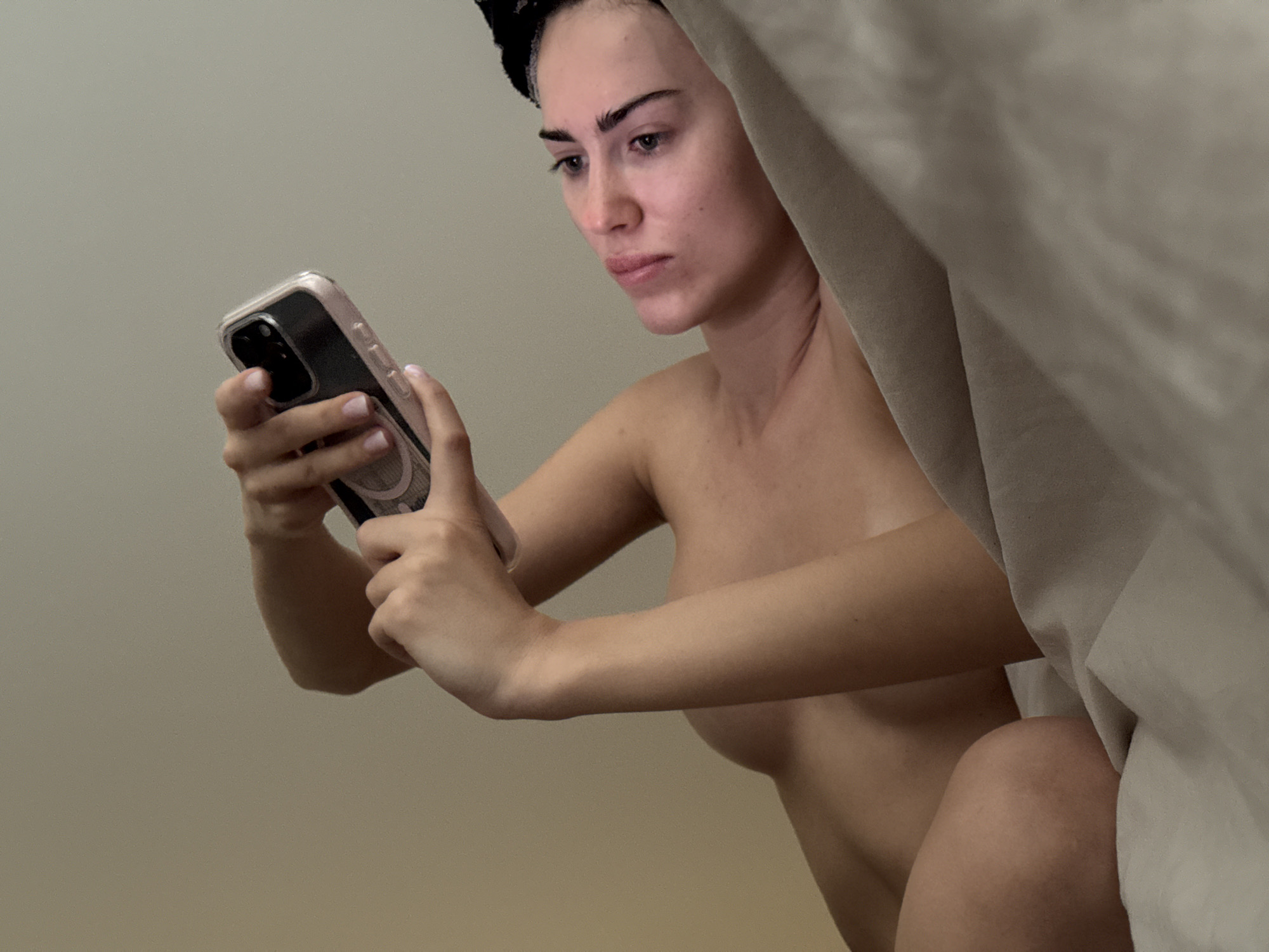 A person with a black head wrap is holding a smartphone in their hands and looking at the screen. The person is positioned to the left of a light-colored drape or sheet. The background is a plain, light-colored wall.