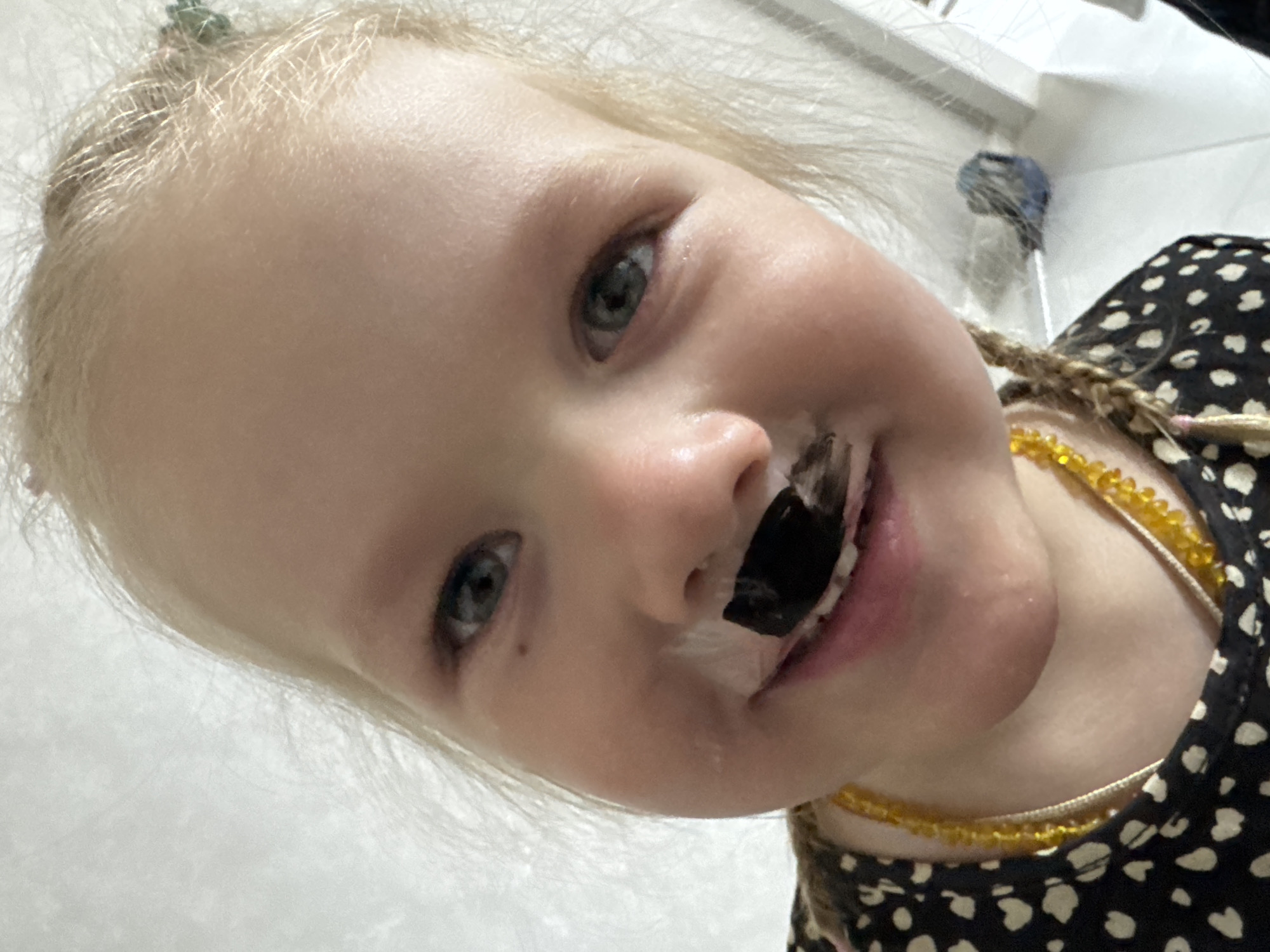 A fair-skinned, blonde-haired child, likely female, is positioned in the foreground. The child is smiling, revealing a dark substance, potentially food or paint, covering their front teeth and upper lip. The child's eyes are light-colored. They are wearing a dark-colored shirt with a light-colored polka dot pattern. A beaded necklace, possibly amber, is visible around the child's neck. The background appears to be a plain, light-colored wall. A small blue object is visible in the upper right corner of the image. The scene is indoors. The specific location within Gornyi (Dvurechenskoe S/P), Russia, cannot be determined from the provided image.