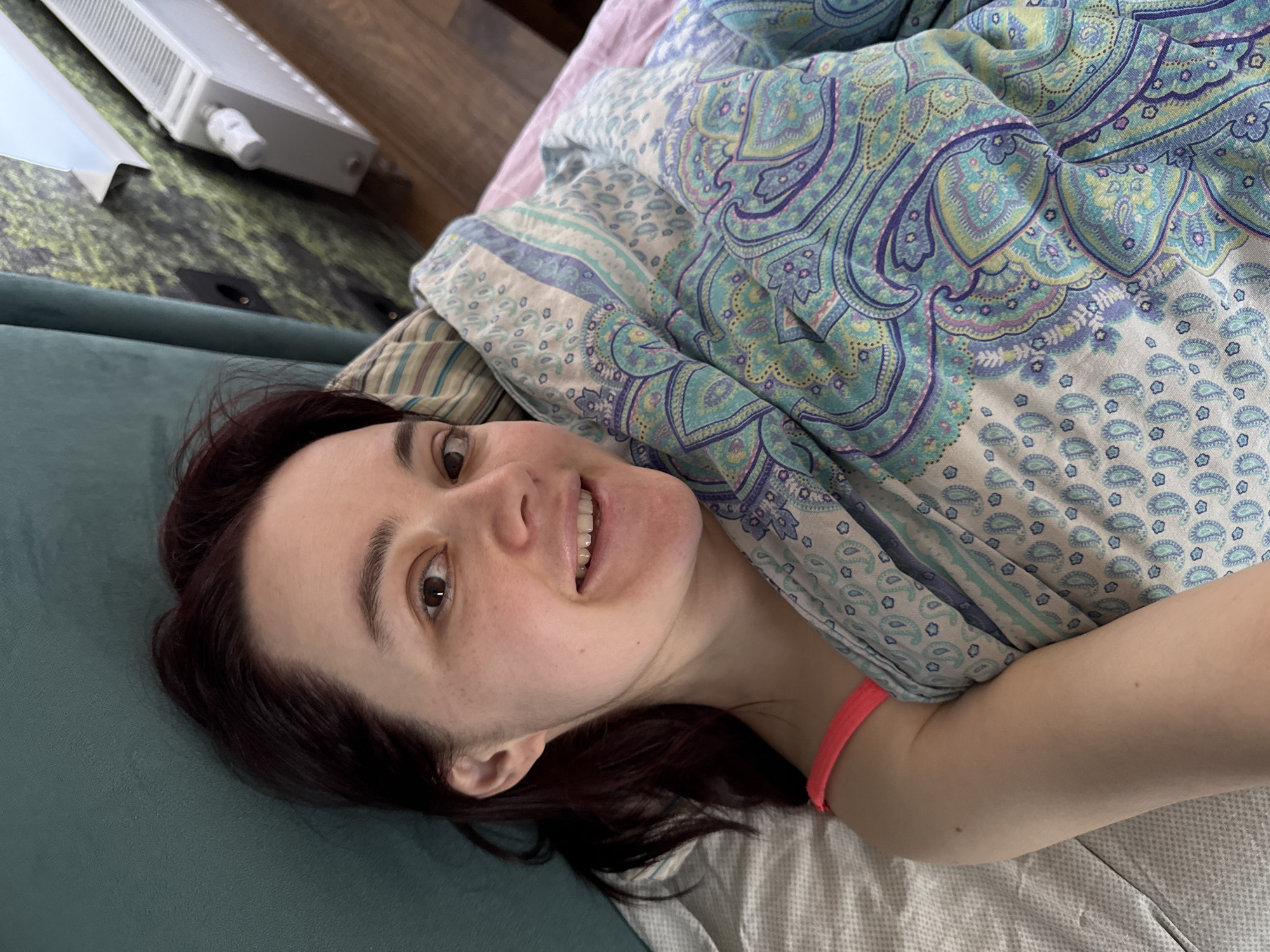 A woman lies on a blue-green couch covered with paisley patterned bedding. She is smiling slightly at the camera. Her hair is dark. A white radiator is visible on the upper left. The location is not identifiable from the image.