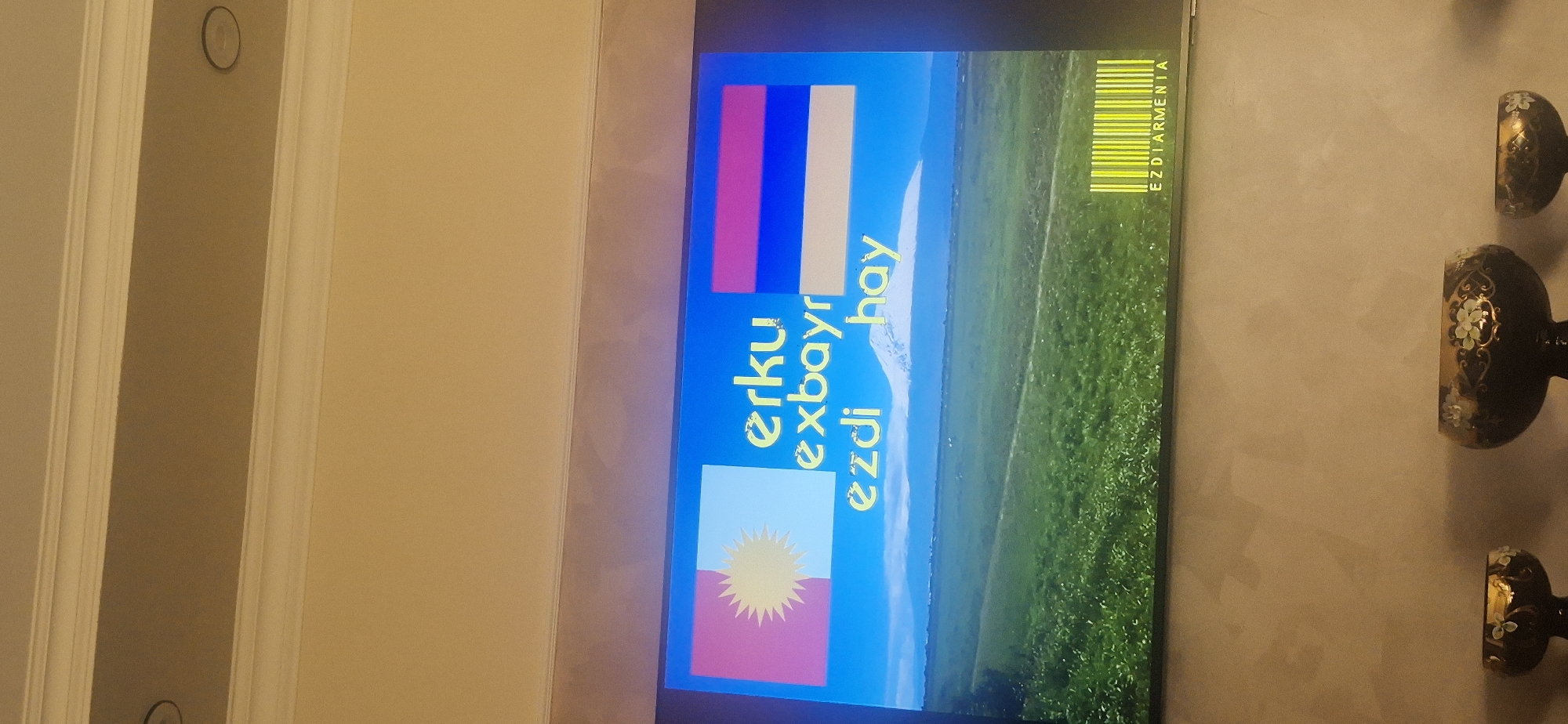 The image shows a television displaying a screen with the flags of Armenia and the Yezidis. Text in yellow spelling out "Erku exbayr" and "hay ezdi" is displayed on the screen. The text "EZDIARMENIA" is also visible, presented as a barcode. A green grassy field is present in the background. The television is located indoors, as evidenced by the adjacent wall decorations and a door frame to the left. The exact city and location cannot be determined from the provided image.