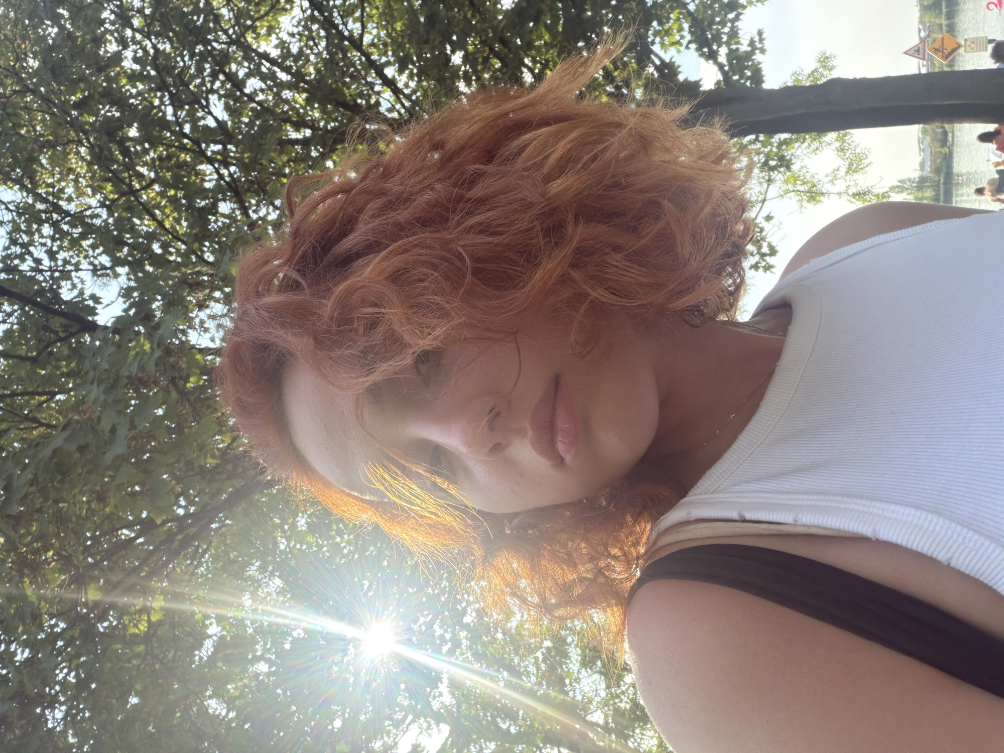 A medium shot, from a low angle, captures a young woman with auburn, curly hair, turned slightly to her left. She wears a white tank top and a black strap is visible over her left shoulder. Bright sunlight streams in from the left, creating lens flares and illuminating her hair and face. She is outdoors, with lush green trees and branches filling the upper portion of the frame against a bright sky. In the background, out of focus, is a body of water with several people visible near the shore. The image appears to have been taken at the Parc de l'Étoile in Illkirch-Graffenstaden, France, given the presence of the lake and the general atmosphere of a park setting.