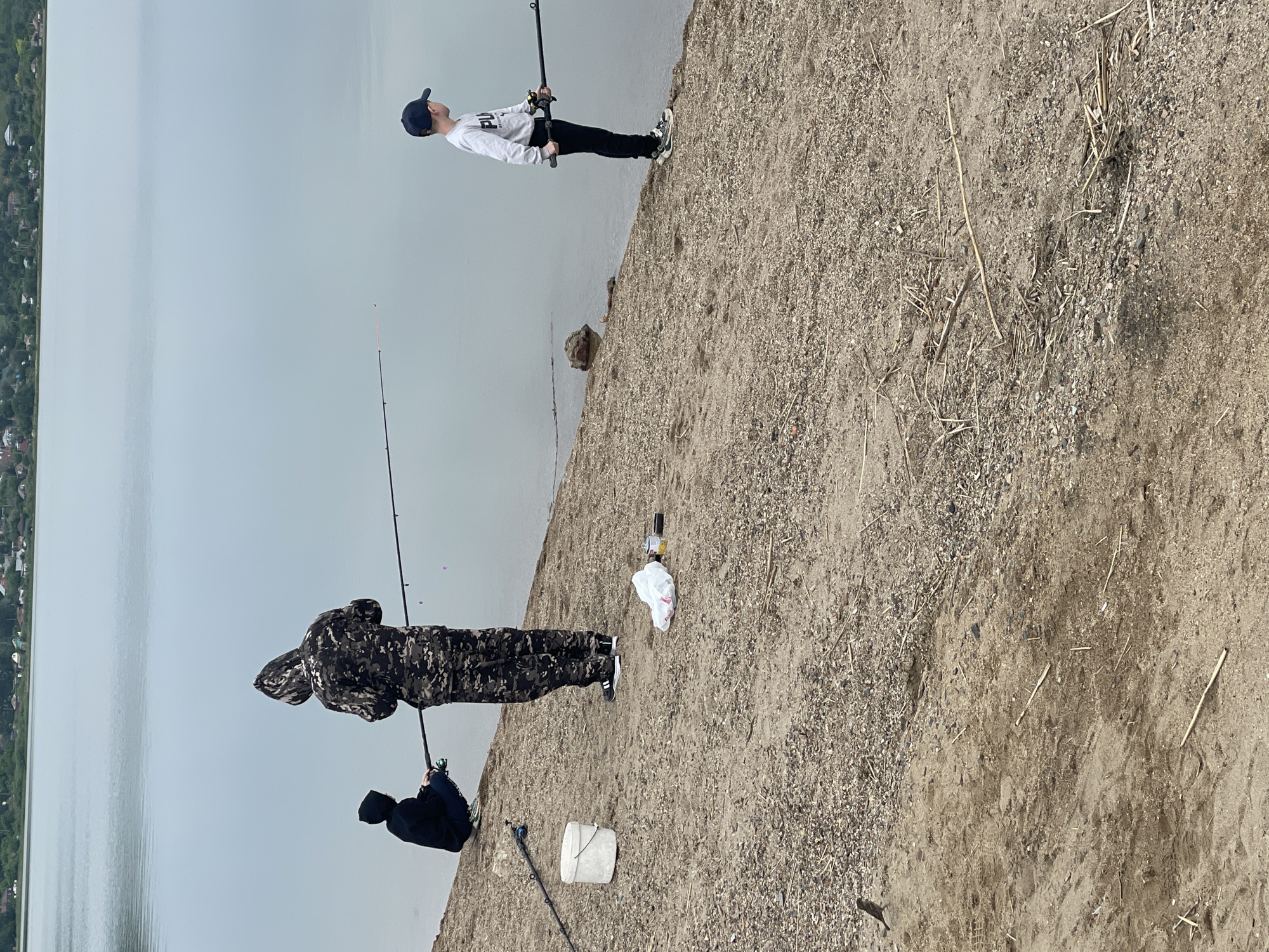 The image shows three individuals fishing along a shoreline in Kokshetau, Kazakhstan. The scene is captured during daylight, with the horizon and a portion of the city visible in the background. The water is a calm, light grey. The fishing takes place on a sandy, grainy ground. The person closest to the camera is wearing a camouflage jacket, while another individual is wearing a dark jacket with a hood. The third fisherman is wearing a white shirt with black pants and a baseball cap. All three are holding fishing rods. A white bucket and plastic bag are placed on the ground nearby.