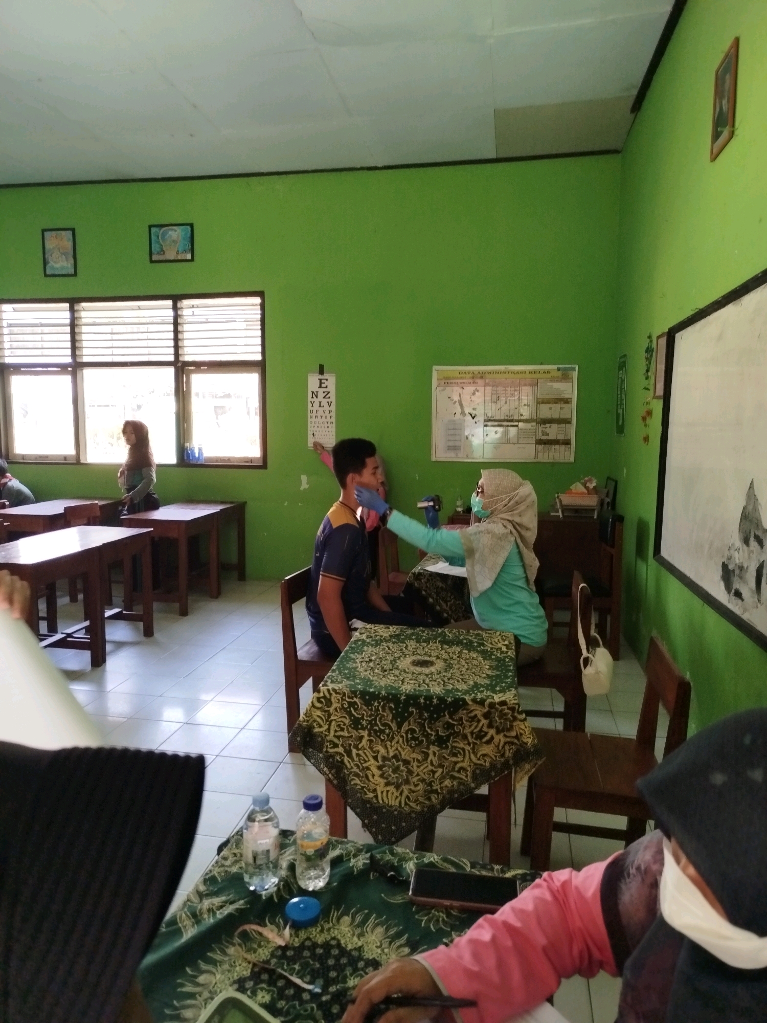 In a brightly lit classroom in Kedungwaru, Indonesia, a medical examination is taking place. The walls are painted a vibrant green, and two framed pictures hang on the left wall. Rows of wooden desks with matching chairs are arranged throughout the room, some covered with patterned cloths.
In the center of the frame, a young man in a blue and yellow striped shirt is seated, appearing to undergo an examination of his mouth by a woman wearing a light blue hijab and a turquoise shirt. She is wearing blue gloves and holding a medical instrument. To her right, a whiteboard with handwritten notes is visible on the green wall, alongside an eye chart.
Near the windows, another woman in a dark headscarf and grey top is standing and looking towards the examination. In the foreground, a person in a pink jacket and black headscarf is seated, partially obscuring the view of a table with two water bottles and a small blue measuring tape. The floor is tiled with a light-colored pattern. The overall atmosphere suggests a school health check-up or a similar medical screening event within an educational setting.