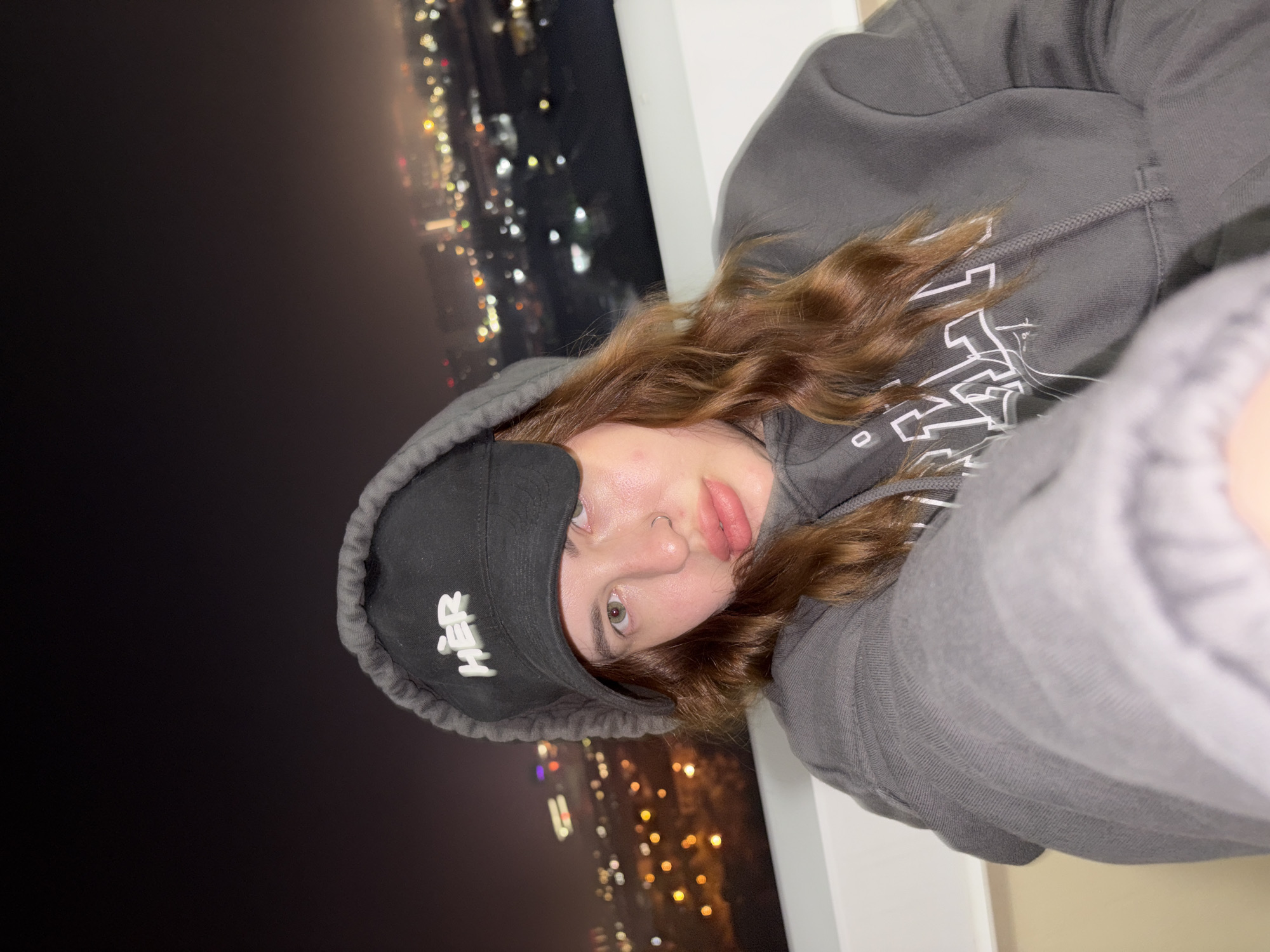 A close-up selfie captures a young person, likely female, tilted to the right, looking directly into the camera. The setting is a nighttime cityscape in Krasnodar, Russia, viewed from a high vantage point, possibly a balcony or window, with blurred, sparkling city lights in the background.
The subject has long, wavy, reddish-brown hair, green/hazel eyes, and wears a septum piercing. Her lips are slightly parted in a subtle pout. She is dressed in a dark grey hooded sweatshirt with the hood pulled up, and a black baseball cap is worn backwards, covering the top of her head. The word "HER" is visible in white block letters on the front of the cap. The grey sweatshirt also features indistinct white graphics or text on its front. A white railing or window frame is partially visible on the right side of the image. The overall impression is a casual, intimate portrait against a vibrant urban night backdrop.