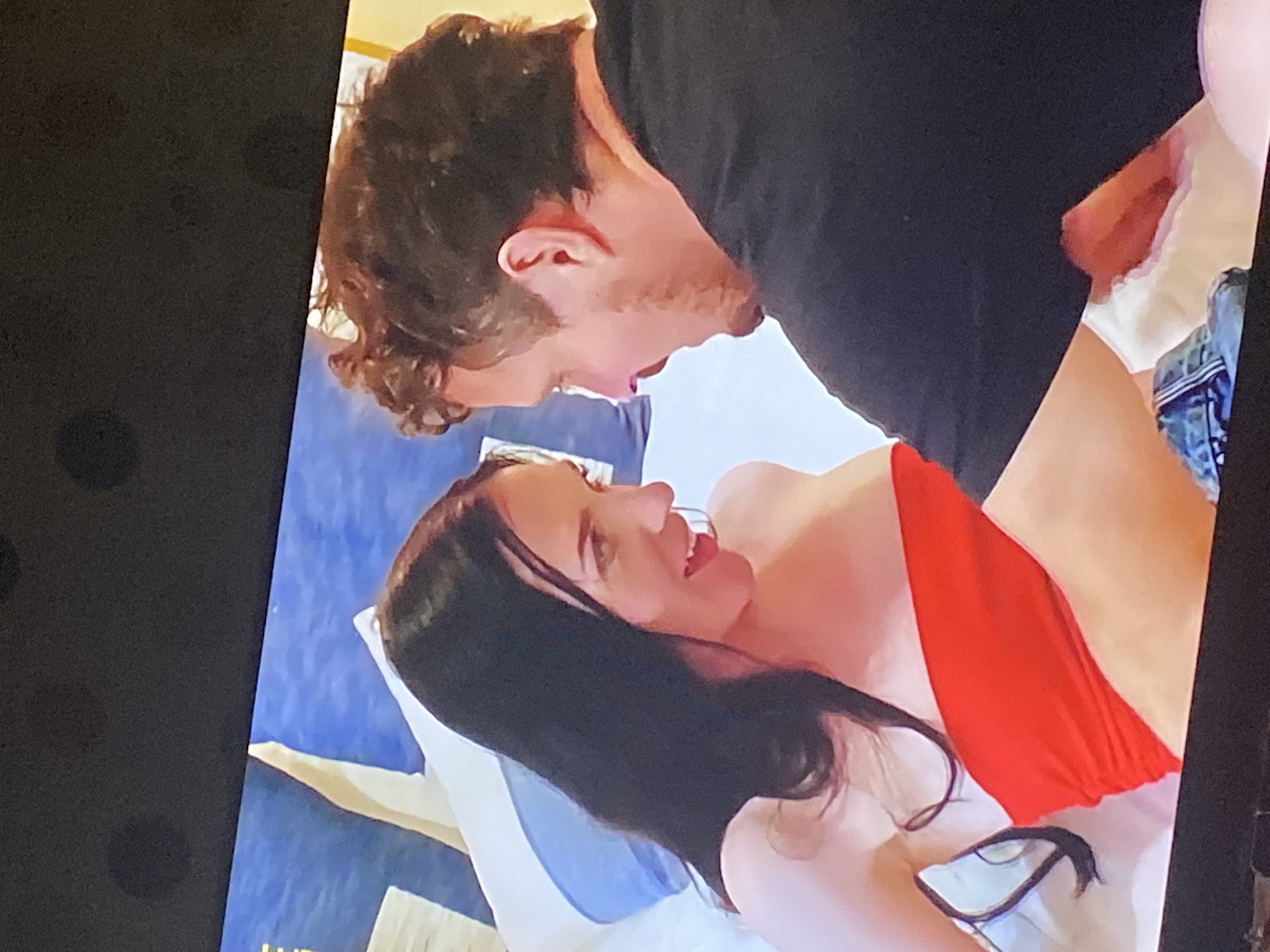 The image shows a close-up of a couple, a woman and a man, looking at each other. The woman has long dark hair and is wearing a red tube top. The man has short, curly brown hair and is wearing a black t-shirt. They are in what appears to be a bedroom, with pillows and bedding visible in the background. The lighting suggests it could be daytime. There is no indication of the city of Brashlen, Bulgaria.