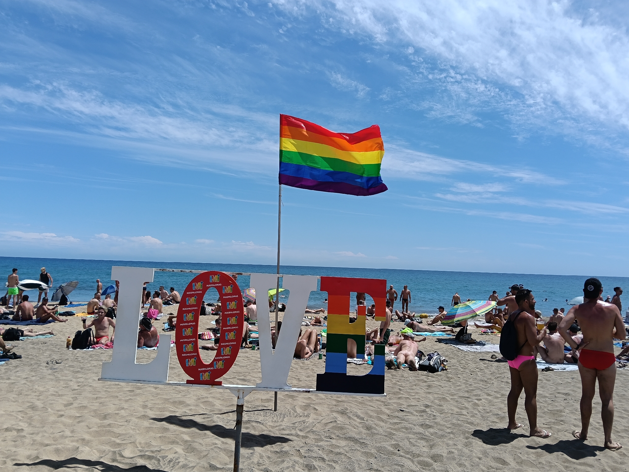 This image shows a beach scene in San Bartolomé de Tirajana, Spain. A large, white "LOVE" sign is positioned on the sand, with the "O" filled with a red logo and the "E" painted in rainbow colors. Behind the sign, a rainbow flag flies on a pole. People are lounging on the beach, some sunbathing and others standing or walking. The ocean is visible in the background, under a partly cloudy blue sky.