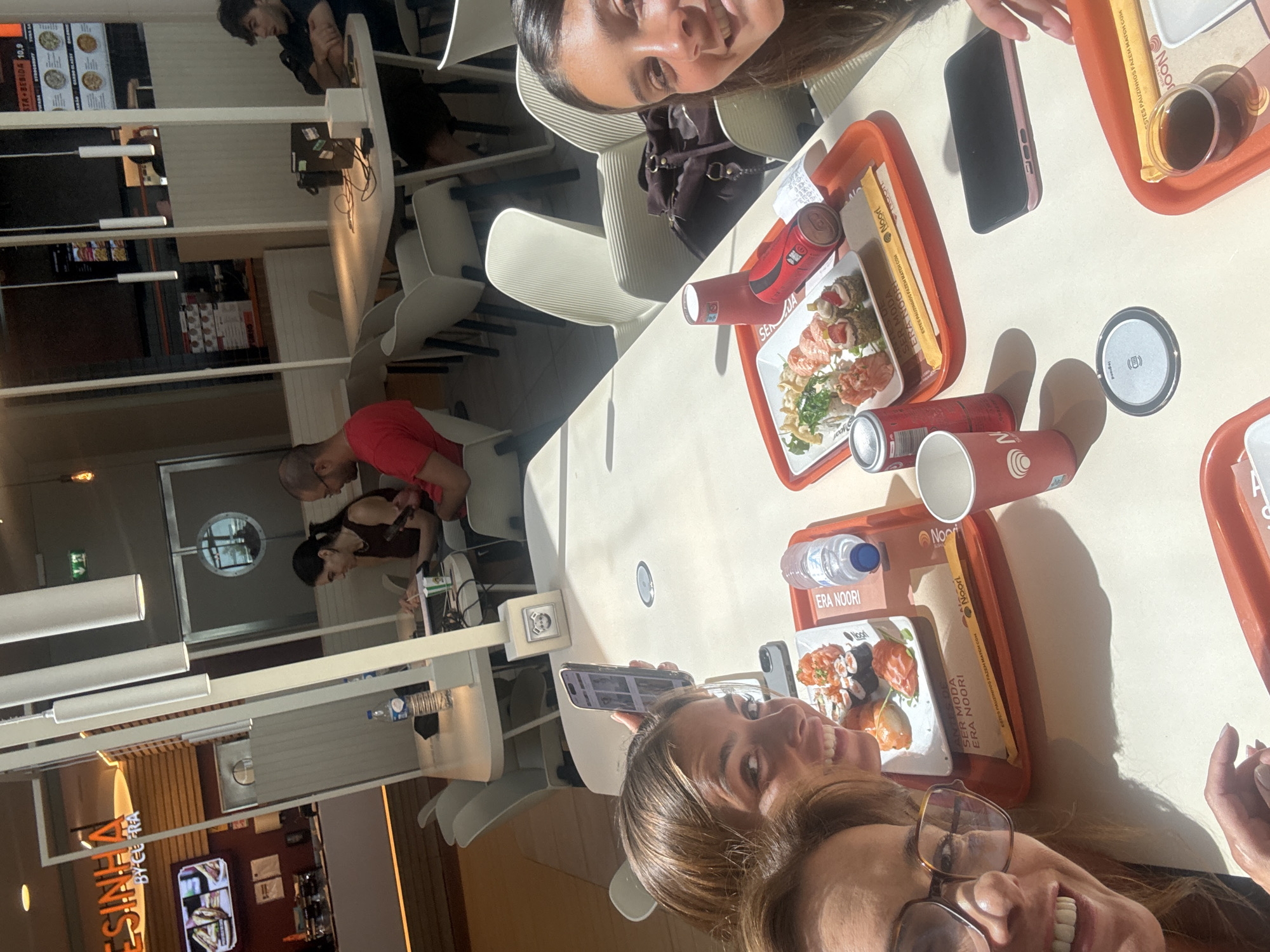 In a bright, indoor setting, likely a restaurant or food court, three young adults are seated around a table, enjoying a meal. The table is laden with orange serving trays containing sushi, chopsticks, drinks, and what appears to be soy sauce. Two of the individuals, a woman and a man, are partially visible in the foreground and mid-ground, engrossed in their phones. Another woman, her face prominently featured in a selfie-style shot, smiles broadly at the camera, her long brown hair framing her face. The scene is illuminated by natural light, casting shadows across the table, suggesting it is daytime. The restaurant appears modern and clean, with rows of chairs and tables visible in the background. The text visible on the trays and packaging indicates a Japanese-inspired cuisine, with brands like "Noori" and phrases such as "SER MODA ERA NOORI" and "ESTES PAUZINHOS FAZEM MATCH COM." The overall mood is casual and convivial, with the individuals appearing relaxed and engaged.