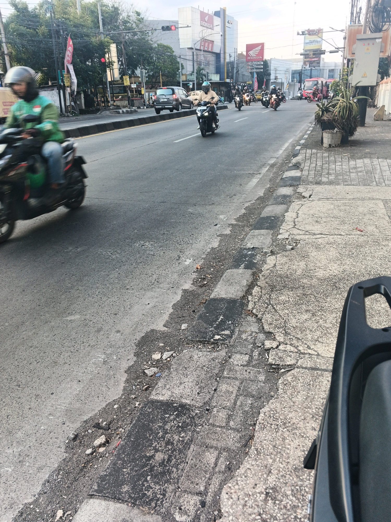 This is a medium-angle, eye-level shot of a street scene in Ungaran, Indonesia, likely captured from the perspective of another motorcycle rider. The focus is on the road and sidewalk, with a procession of motorcycles moving along the street in the background. The road appears to be asphalt, with some visible damage and debris along the edge. A concrete sidewalk runs parallel to the road, also showing signs of wear and tear.
In the immediate foreground, the back of a dark-colored motorcycle or scooter is visible, suggesting the photographer is on a similar vehicle. To the left, a motorcyclist in a green jacket and helmet is in motion, with the vehicle blurred due to movement. Further down the road, several other motorcycles are also in motion, some carrying multiple riders. One rider, in a light-colored shirt and helmet, is clearly visible in the middle ground.
The background features a city street with various buildings, including those with "HONDA" signage, indicating commercial establishments. Traffic lights are visible, with one showing a red light. Trees line the side of the road, and power lines and utility poles are present. The overall impression is of a busy urban environment with active motorcycle traffic. The lighting suggests it is daytime, possibly late afternoon, with a bright sky.
