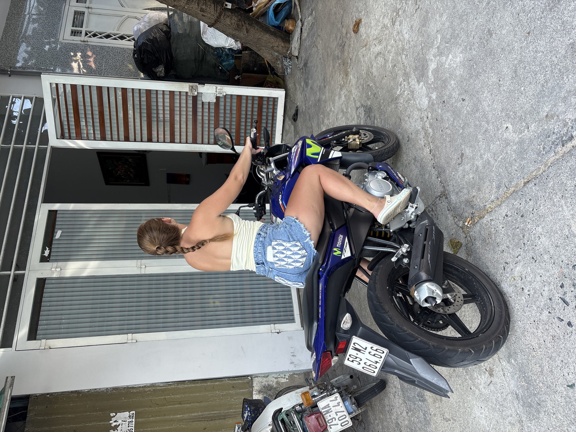 A full shot depicts a woman sitting on a motorcycle in Nha Trang Ward, Vietnam. The woman, facing away from the camera, has blonde hair braided down her back. She wears a white tank top and denim shorts with a pattern of fish on the back pockets. The blue and black motorcycle is parked on a concrete surface, with a license plate reading "59-M2 064.66". Behind the motorcycle and to the left of the woman, part of a white motorcycle is visible, also with a license plate. The background includes a building with a white door and windows. Above the door, there is a dark painting hanging on the wall. A tree is present in the top right corner of the image. Trash bags are positioned near the tree.