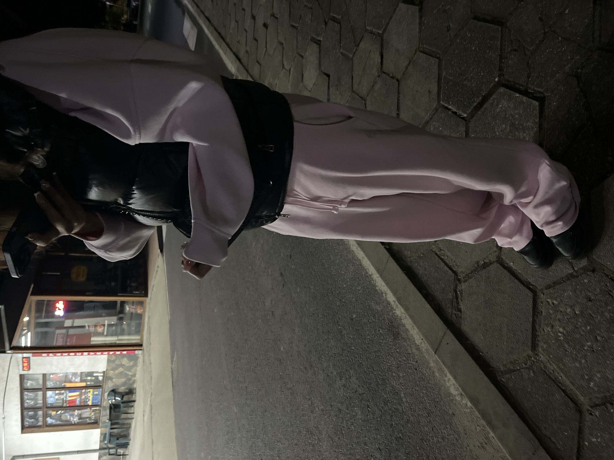 A female figure is standing on a sidewalk in Zlatograd, Bulgaria, based on the Cyrillic text visible on a nearby store. She is wearing a light pink tracksuit with a black puffer vest. The ground is paved with concrete slabs on one side and a smoother, asphalt-like surface on the other. She is holding a mobile phone. The background features a street with a store front.