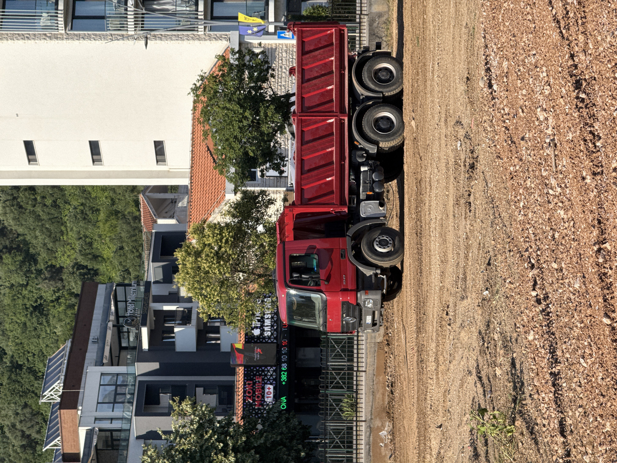 A red dump truck is parked on a dirt road next to a building complex in Budva, Montenegro. The building features modern architecture with glass balconies and solar panels. Signs on the building indicate it is "Hotel Millennium SPA& WELLNESS" and "ZOKI MOBILE" with a phone number. The truck has a large red cargo bed and visible tires. The scene appears to be an outdoor setting under daylight, suggesting a clear day. The surrounding area is a mix of urban development and natural greenery with trees on a hillside in the background. The dirt road shows tire tracks, indicating recent vehicle activity. The overall impression is a snapshot of a construction or delivery scene in a coastal Montenegrin town.