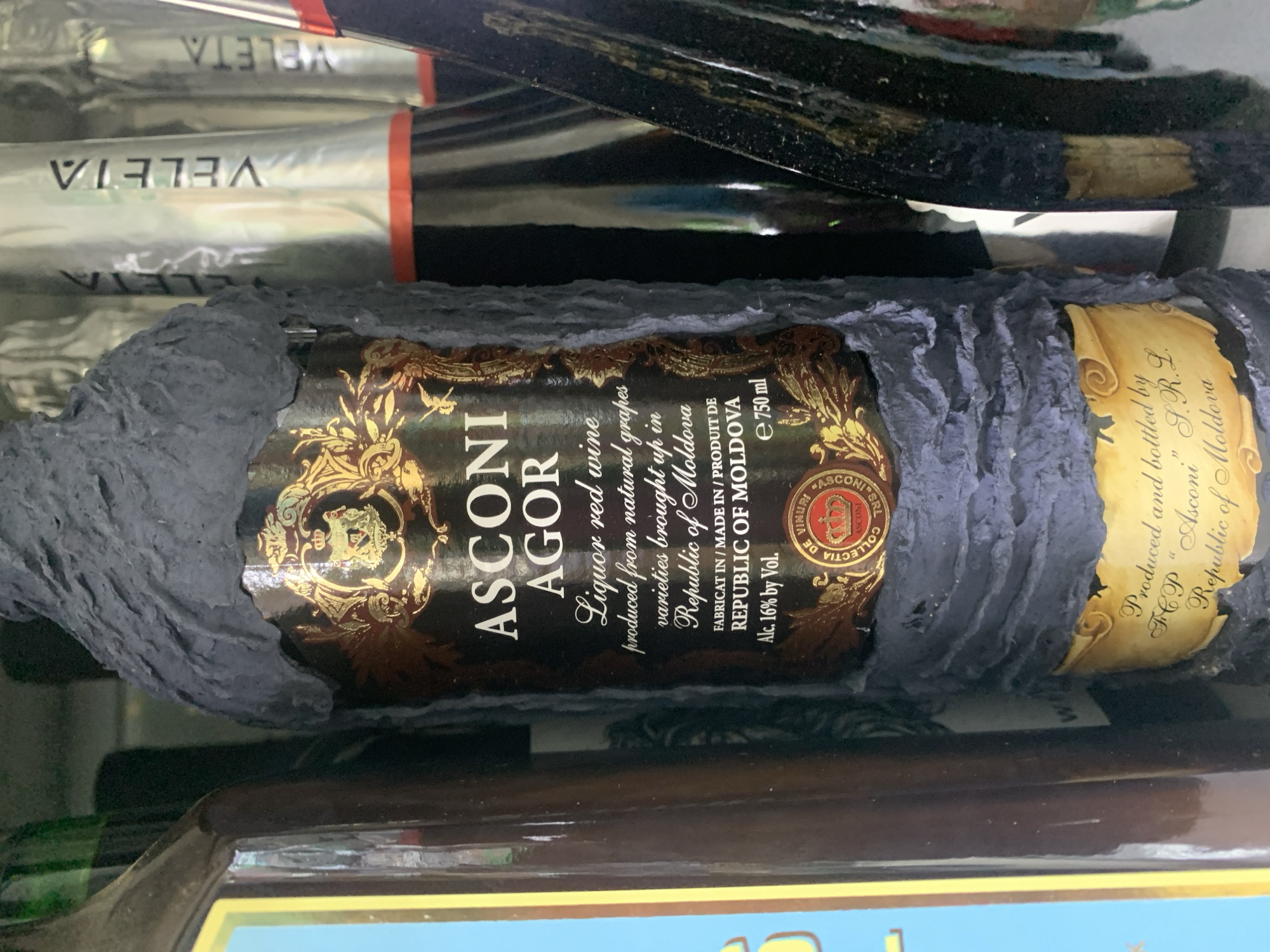The image shows a close-up of a bottle of "ASCONI AGOR" liquor red wine. The bottle is dark and appears to be partially covered in a textured gray material. A label on the bottle has gold embellishments and text that includes "ASCONI AGOR," "Liquor red wine," "produced from natural grapes varieties brought up in Republic of Moldova," "FABRICAT IN / MADE IN / PRODUIT DE REPUBLIC OF MOLDOVA," and "Alc. 16% by Vol." Below this text is a circular seal with a crown and the text "COLLECTIA DE VINURI," "ASCONI," and "SRL." Further down the bottle, a yellow scroll-like label has the text "Produced and bottled by FCP "Asconi" S.R.L. Republic of Moldova." The bottle is positioned among other bottles, some of which have silver foil caps and are labeled "VELETA." The lighting suggests an indoor setting, possibly a shelf in a store or cellar. The scene is static, with no people or actions depicted.