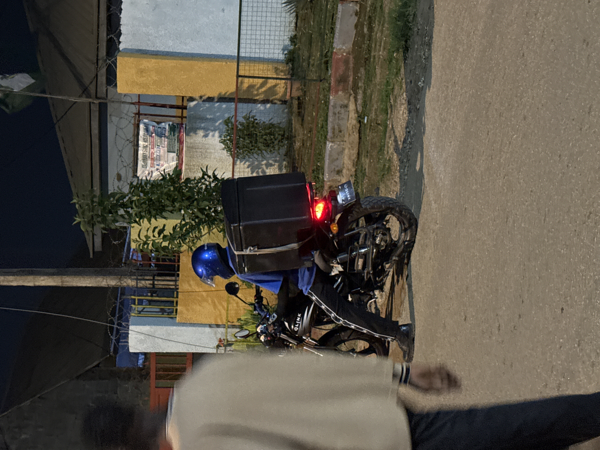 A person wearing a blue helmet and a blue shirt is sitting on a black motorcycle. The motorcycle has a large black storage box mounted on the back. The person's pants have a white stripe pattern. The motorcycle has the word "GLINK" written on its side. The scene appears to be outdoors at night, with a building and a fence in the background. There is also a person in the foreground on the left side of the image, partially out of focus and facing away from the camera. The ground is paved. The image is taken in Abuja, Nigeria.