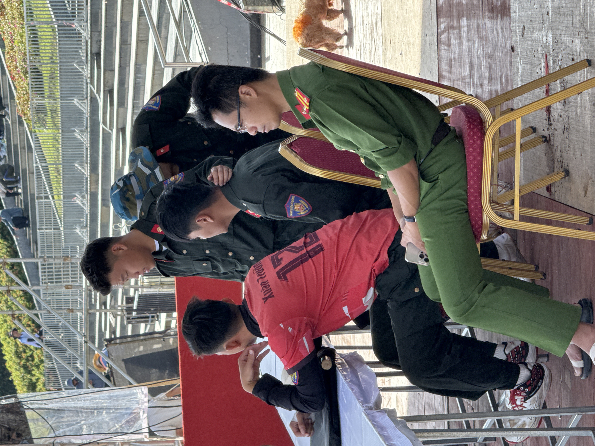 Four men in police uniforms are seated or standing near a red barrier in what appears to be an outdoor public gathering area in Da Lat, Vietnam. The man on the far right is wearing a green uniform with glasses, looking to the side. The man next to him, to his left, is wearing a red shirt with black sleeves and the number "12" on the back. He is leaning on his hand, looking down. Two other men in dark uniforms are standing behind them, one in the center with a camouflage hat and the other on the far left. In the background, there are tiered seating areas and scaffolding, suggesting a venue for an event. A small dog is visible in the background to the right. The ground is a wet, wooden surface.