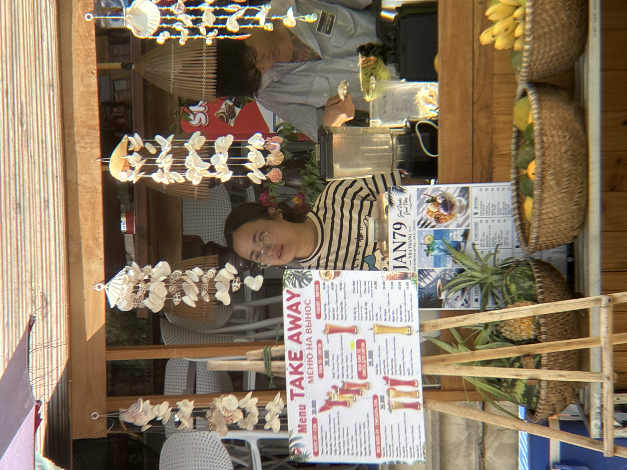The image depicts a roadside vendor stall in Nha Trang, Vietnam. A woman wearing glasses and a striped shirt stands behind a "TAKE AWAY MENU" sign, which lists various drinks and their prices in Vietnamese Dong. To her right, a person in a light blue striped shirt appears to be preparing drinks, with a blender and various containers visible. In front of the stall, woven baskets hold fresh fruits, including watermelons, pineapples, bananas, and avocados. The stall is decorated with shell wind chimes. The overall scene suggests a casual outdoor food and beverage establishment in a tropical setting.