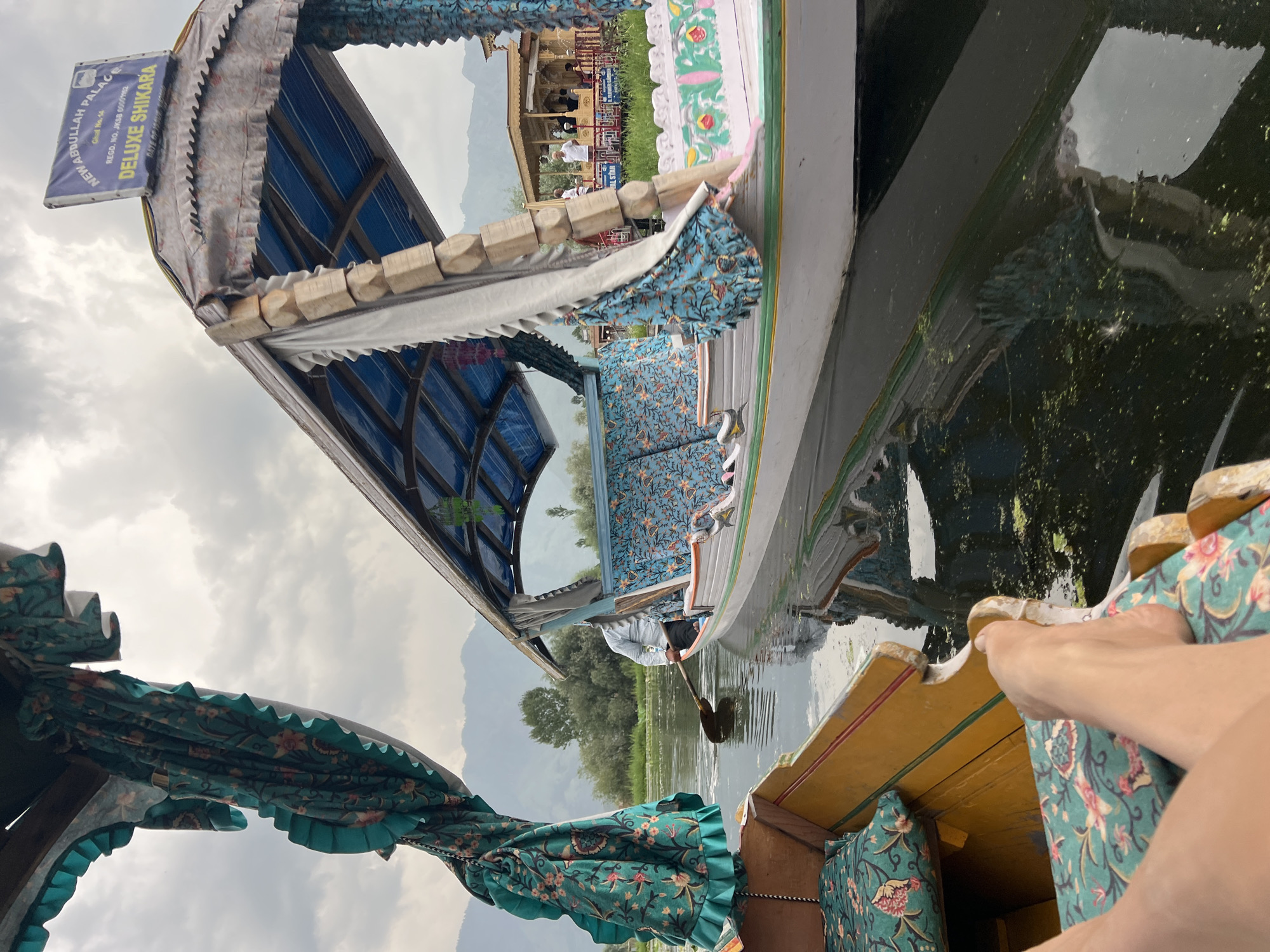 This image captures a serene scene on the Dal Lake in Srinagar, India. Two ornate Shikaras, traditional Kashmiri boats, are prominently featured. The foreground shows a person's arm and hand resting on the brightly painted wooden interior of a Shikara, upholstered in a vibrant floral fabric. The boat's canopy is adorned with a similar decorative textile.
In the middle ground, another Shikara is visible, propelled by a person using a paddle. This boat also boasts colorful upholstery and a patterned canopy. The backdrop reveals a glimpse of the shore with traditional Kashmiri architecture, possibly a palace or houseboats, against a backdrop of lush green trees and distant, hazy mountains. The sky is overcast, suggesting a cloudy or potentially rainy day.
A sign on the first Shikara reads "NEW ABDULLAH PALACE REG D. NO. JKSB 00007487 DELUXE SHIKARA". The overall mood is peaceful and idyllic, highlighting the unique cultural experience of a Shikara ride on the famous Dal Lake.