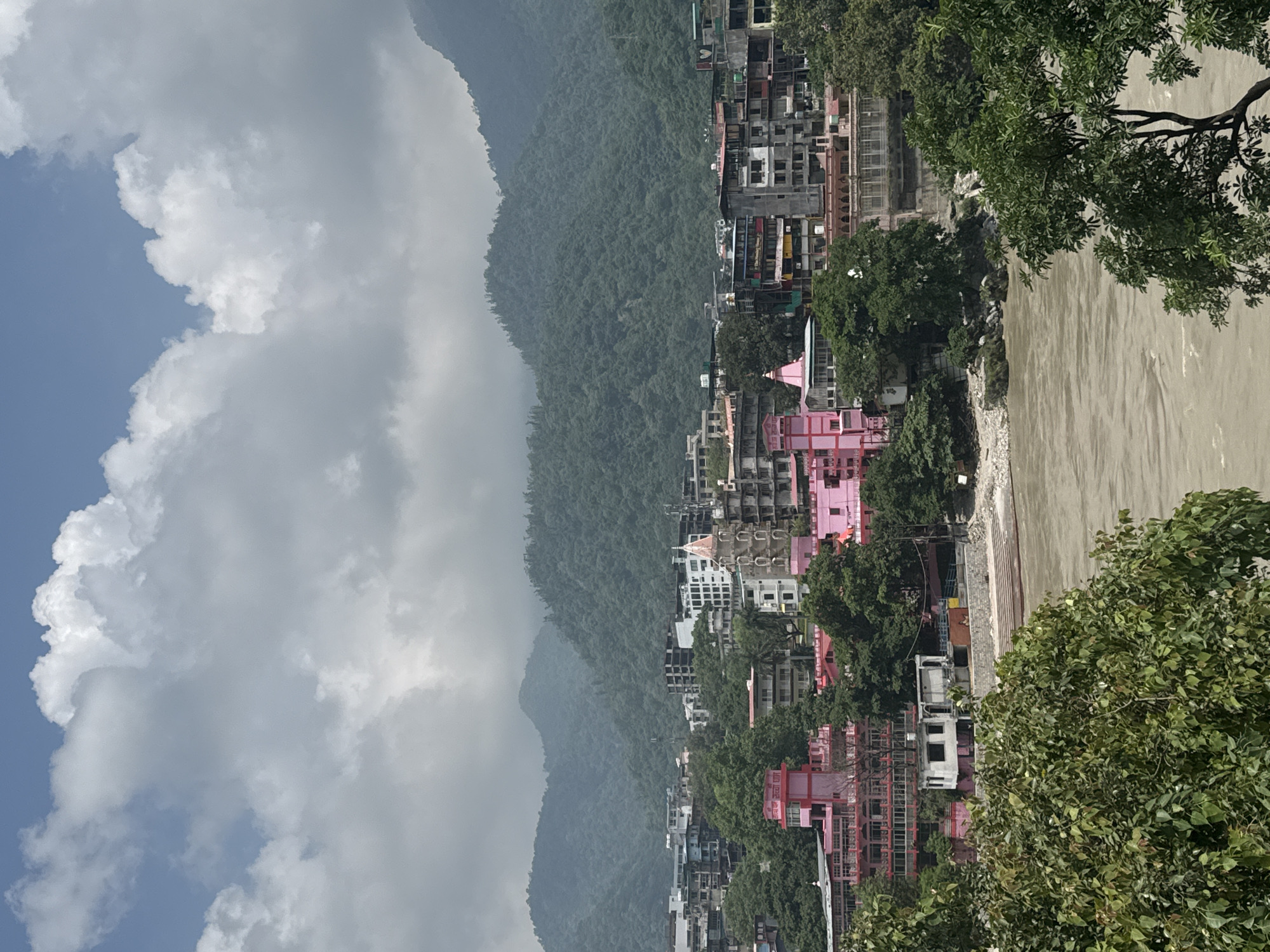 The image depicts a panoramic view of Rishikesh, India, with the Ganges River flowing prominently in the foreground. The river's murky brown water is visible, with visible currents and ripples. Along the riverbank, there are stone steps leading down to the water, suggesting a ghat.
The midground of the image showcases the cityscape of Rishikesh. Numerous buildings of varying heights and architectural styles are present, with a dominant palette of pink and white structures interspersed with more modern concrete and glass buildings. A distinctive pink tower with a spire, possibly a temple or a significant landmark, stands out amongst the other buildings. Lush green trees are scattered throughout the urban landscape, softening the concrete structures.
In the background, imposing mountains covered in dense green vegetation rise majestically. The sky above is partially obscured by large, fluffy white clouds, hinting at a partly cloudy day. The overall scene conveys a sense of a bustling town nestled within a beautiful natural landscape.