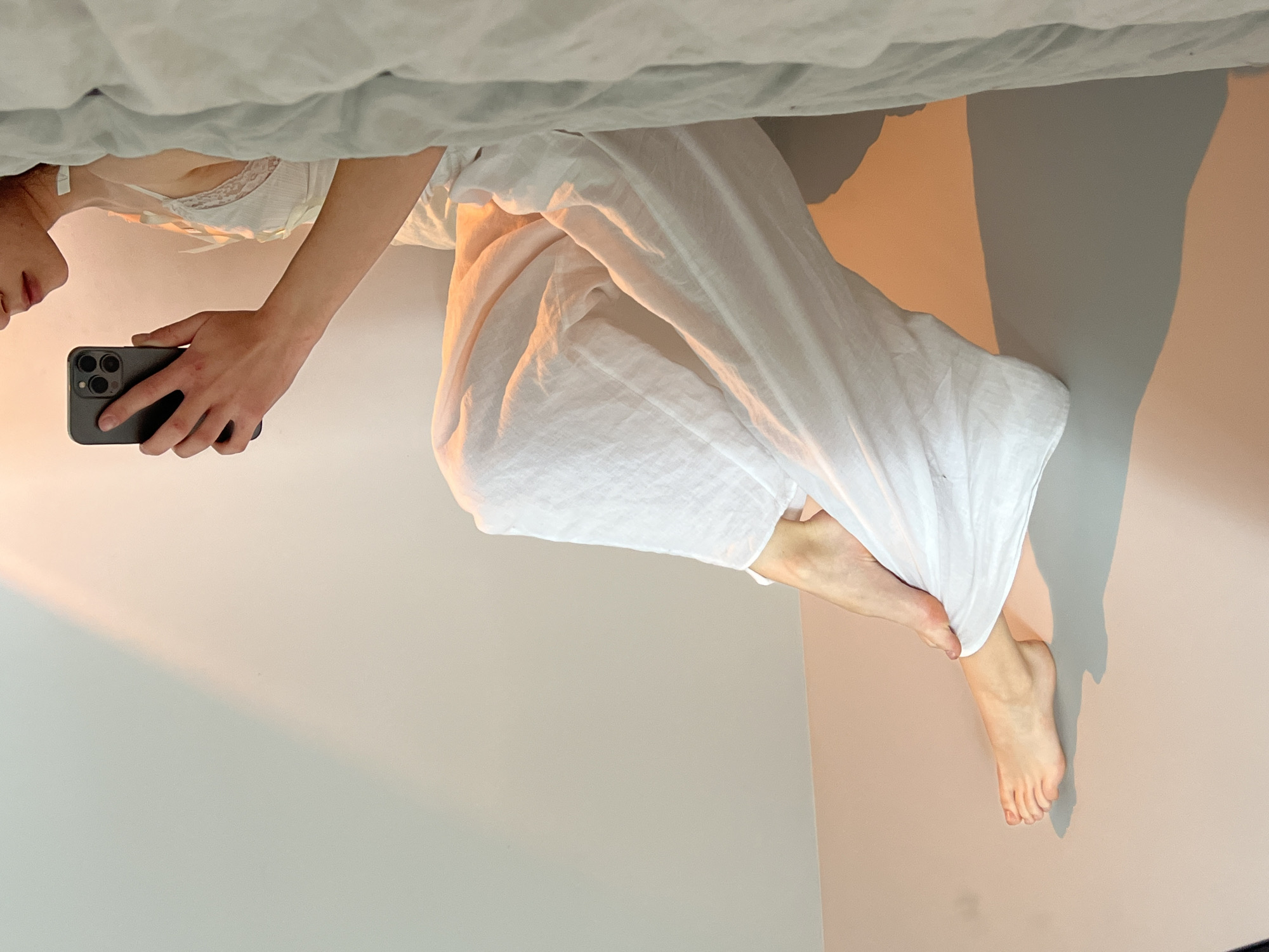 A young woman is lying on a bed, dressed in white linen pants and a white top with a lace trim. She is holding a dark-colored smartphone in her hand, presumably taking a photograph. The background is a plain, light-colored wall. There are no identifiable landmarks or features to place the scene within the city of Ternopil, Ukraine.