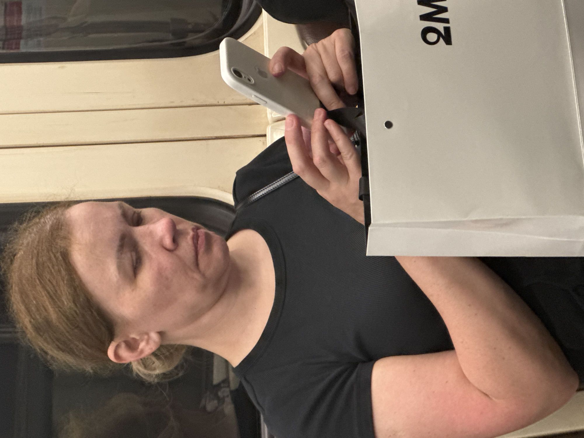 The image shows a woman with light brown hair pulled back in a bun, wearing a black t-shirt. She is seated on what appears to be public transportation, possibly a train or bus, indicated by the beige paneling and window reflections typical of Moscow's metro system. The woman is looking down at her white smartphone, which she holds in her hands. In front of her is a white shopping bag with the logo "2M" printed in black. The lighting suggests an indoor environment, possibly during the day, with the reflections in the background hinting at a moving vehicle. The overall scene depicts a common urban moment of a passenger using their phone while commuting.